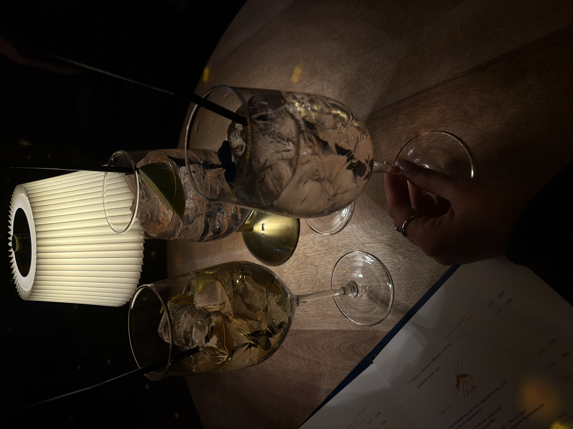 The image shows a table setting in what appears to be a dimly lit restaurant or bar. There are three wine glasses filled with ice and liquid, possibly cocktails, with straws. A lamp with a pleated lampshade provides some light. A hand with a ring is visible, possibly holding a wine glass. A menu is placed on the table, containing text in Russian, including the names of dishes (Borsch, Mushroom cream-soup, Potato gnocchi with cured duck). Based on the text and overall style, the location is likely in Russia. The OCR text indicates the city as Красноярск (Krasnoyarsk).