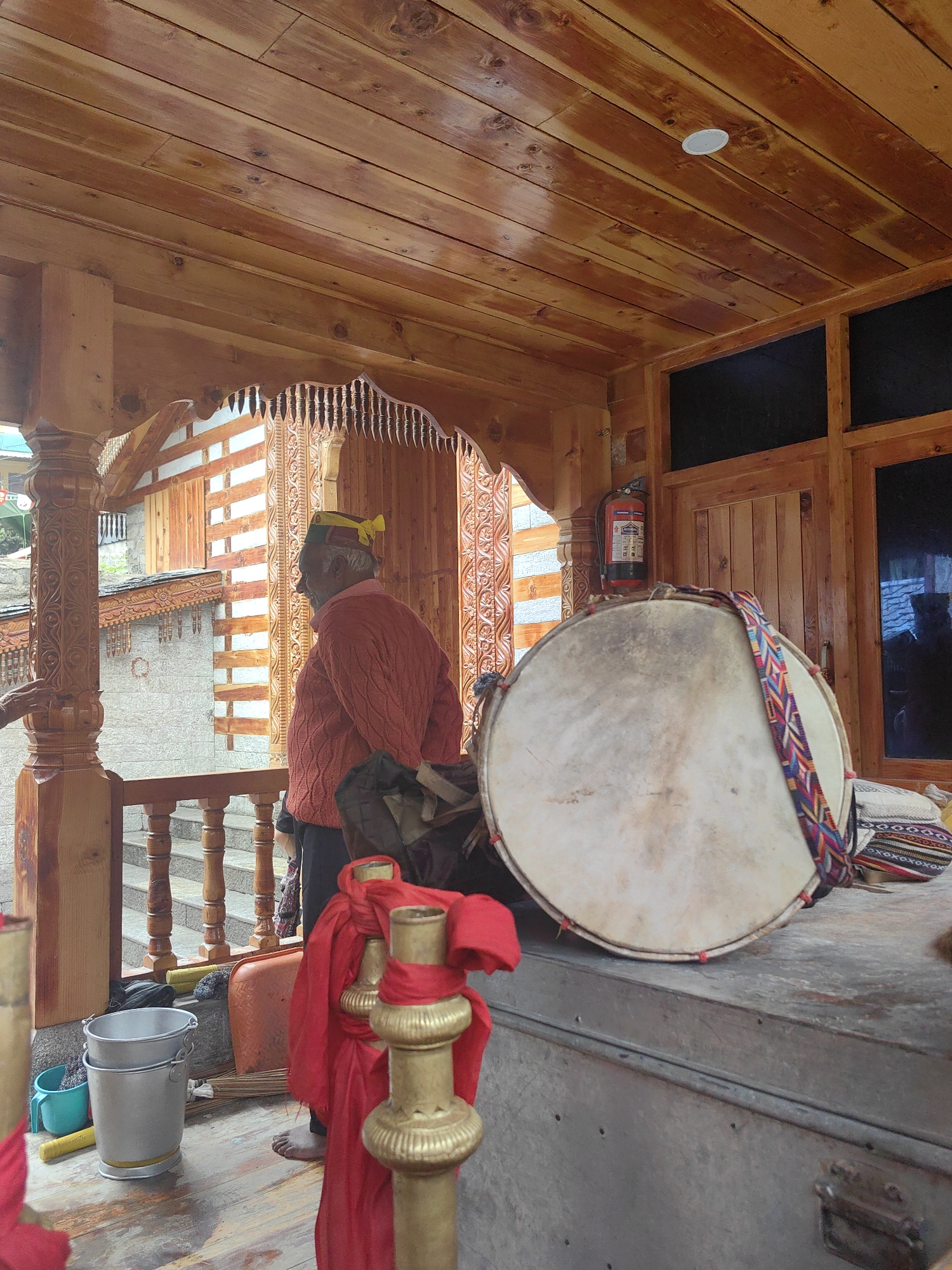 A man in a red knitted sweater and a yellow and green headscarf stands on a wooden porch, facing away from the camera. He is positioned to the left of a large, round drum with a colorful woven strap. The drum rests on a metal surface, possibly a trunk. To the left of the man, stacked metal buckets and a blue plastic container are visible on the wooden floor. Red fabric is draped over decorative golden posts. In the background, a stone building with wooden accents and stairs can be seen. A fire extinguisher is mounted on a wooden wall to the right of the man. The porch features intricately carved wooden pillars and a slatted wooden ceiling. The scene appears to be in Vashisht, India, given the architectural style and the man's attire.