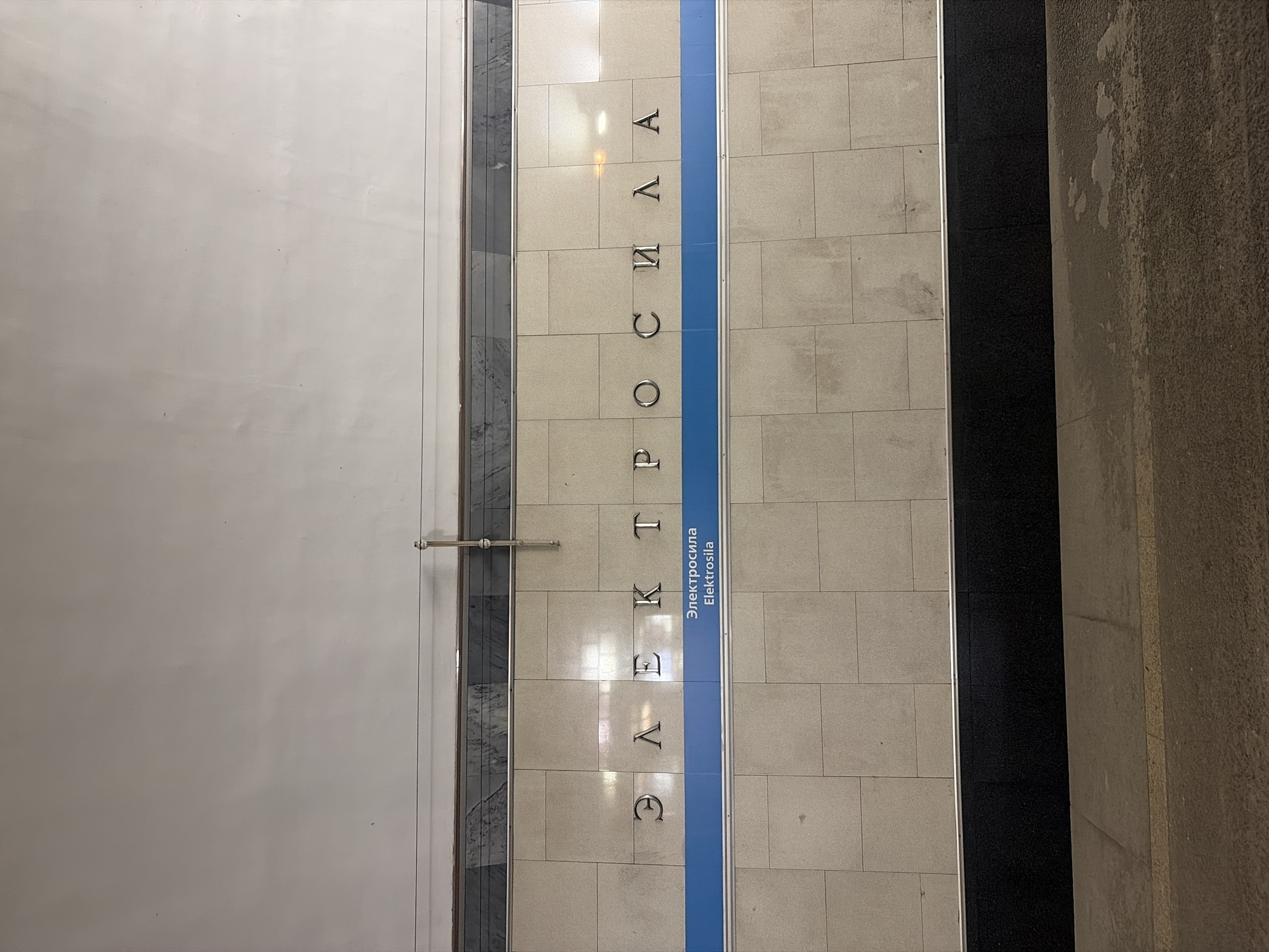 The image depicts a section of the Elektrosila metro station wall in Saint Petersburg, Russia. The wall is comprised of square, beige-colored tiles with blue vertical stripe. The station's name "Elektrosila" is spelled out vertically in both Cyrillic ("Электросила") and Latin alphabets. To the left of the tiled wall is a smooth white surface and a metal bar. To the right is a dark strip, and a dirty stone column.