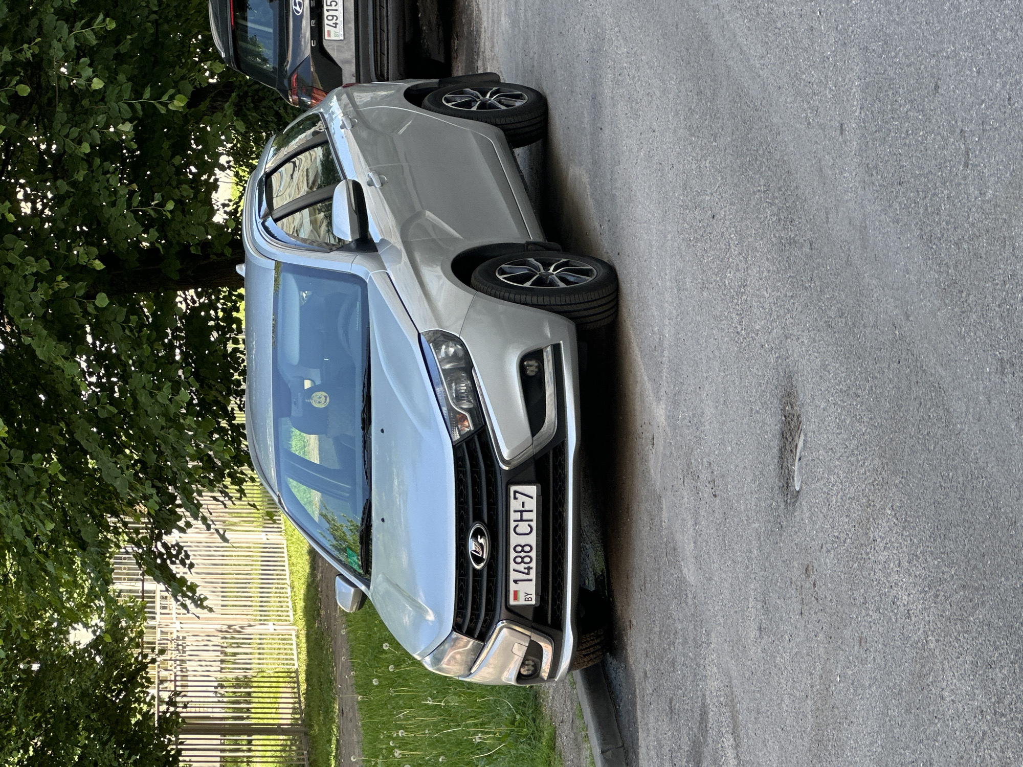 A silver Lada Vesta wagon, license plate BY 1488 CH-7, is parked on a street in Minsk, Belarus. The car is facing the viewer and is slightly angled to the left. The front of the car is visible, including the headlights, grille, and license plate. The car has tinted windows and alloy wheels. In the background, a black Hyundai Creta is parked behind the Lada. A white metal fence and lush green trees are visible behind the cars. The street is paved with asphalt, and there is a patch of grass and dandelions to the left of the car. The scene appears to be a residential area, likely in a city or suburban environment, given the presence of parked cars and residential fencing.