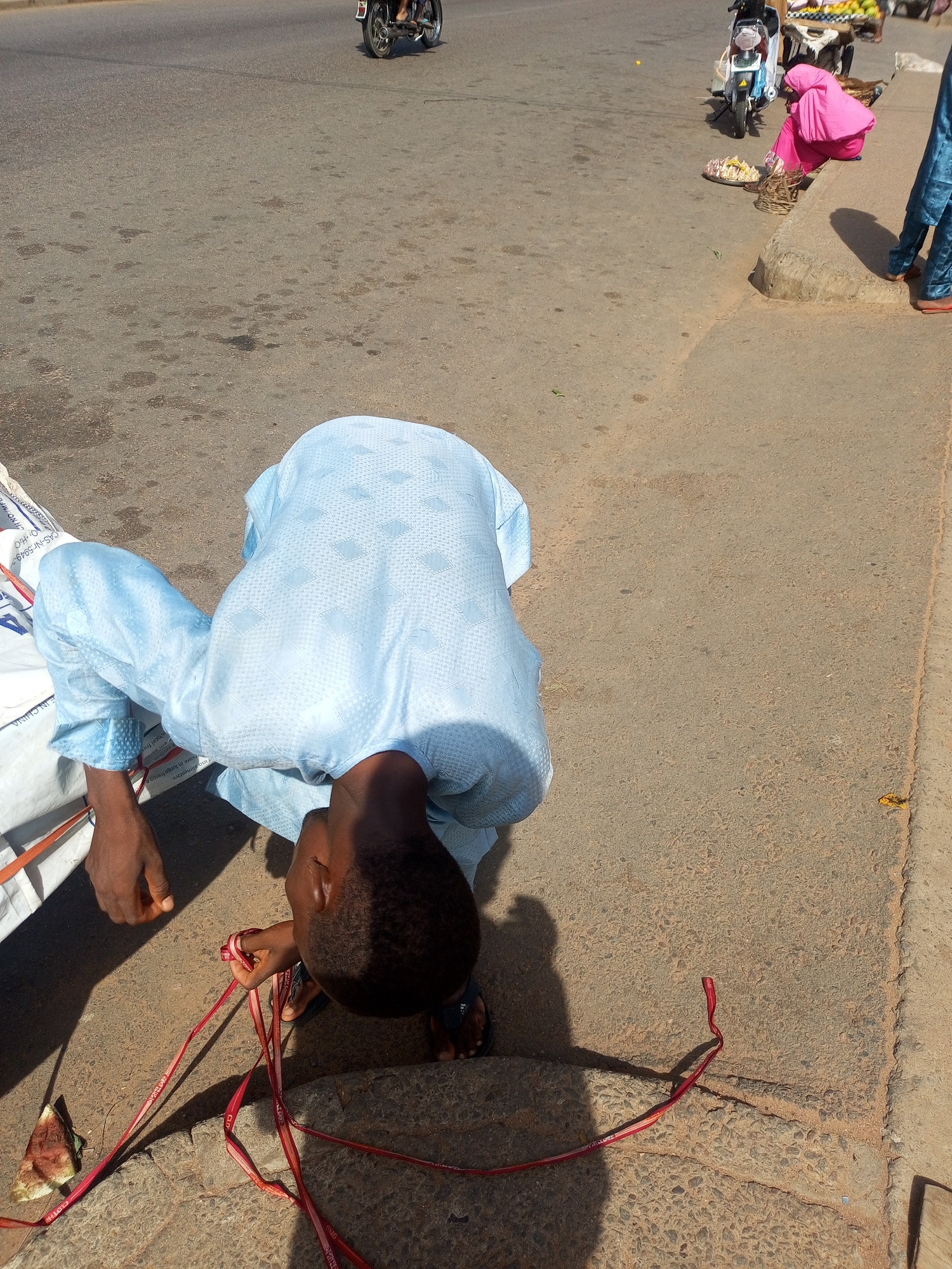 This media file depicts a bustling street scene in Tempure, Nigeria, bathed in bright daylight with strong, clear shadows. In the foreground, a young man, dressed in a light blue traditional garment with a subtle diamond pattern, is bent over with his head down, seemingly focused on the ground. He appears to be untangling or manipulating a collection of red ribbons or straps, which are scattered on the concrete edge of the sidewalk. A discarded piece of watermelon rind lies nearby. To his left, several large white sacks are visible, one bearing text such as "FIN CHINA," "CAS-Nr:5949-," "07 H2O," and "duso alimentare." The red ribbons also show repeating text like "COT" and "COTI8."
Further back in the mid-ground, on the right side of the frame, another individual wearing a vibrant pink garment is seated on the raised concrete curb, next to a parked motor scooter. In front of them, a tray holds a pile of light-colored snacks or food items. The legs of a third person, dressed in blue trousers and sandals, are partially visible on the far right. In the distance, on the asphalt road, a motorcycle is seen. The overall scene suggests a typical roadside environment with daily activities, possibly commerce, taking place under sunny weather conditions.