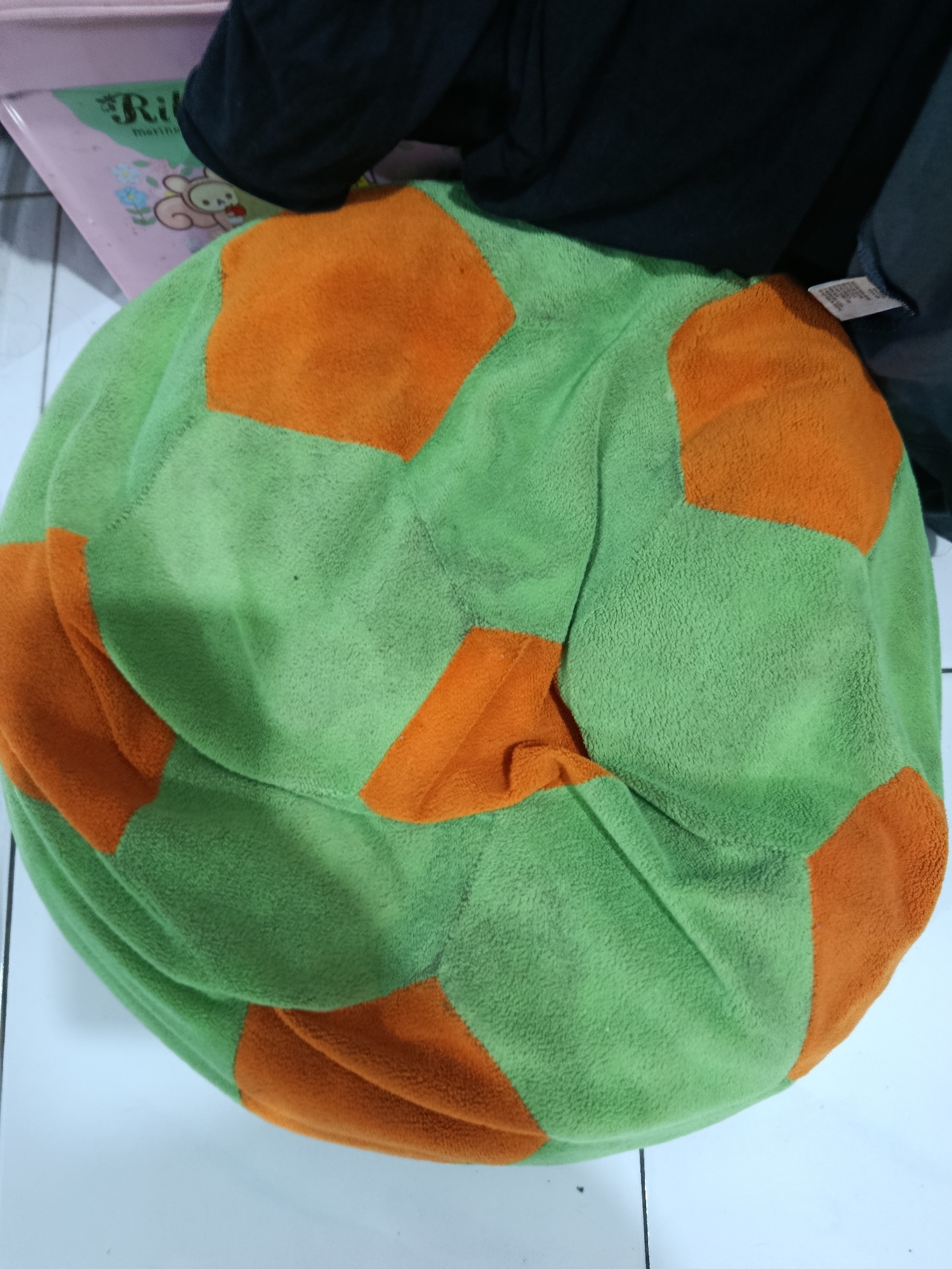 A large, round, plush toy resembling a soccer ball, with alternating green and orange hexagonal panels, is lying on a white tiled floor. A portion of a pink plastic object with a drawing of a cartoon bear and flowers is visible in the upper left corner. Dark fabric, likely clothing, is draped over the toy and extends to the upper right. A white clothing tag is attached to the dark fabric in the upper right. The scene is indoors and appears to be in a residential setting in Wanasari, Indonesia.