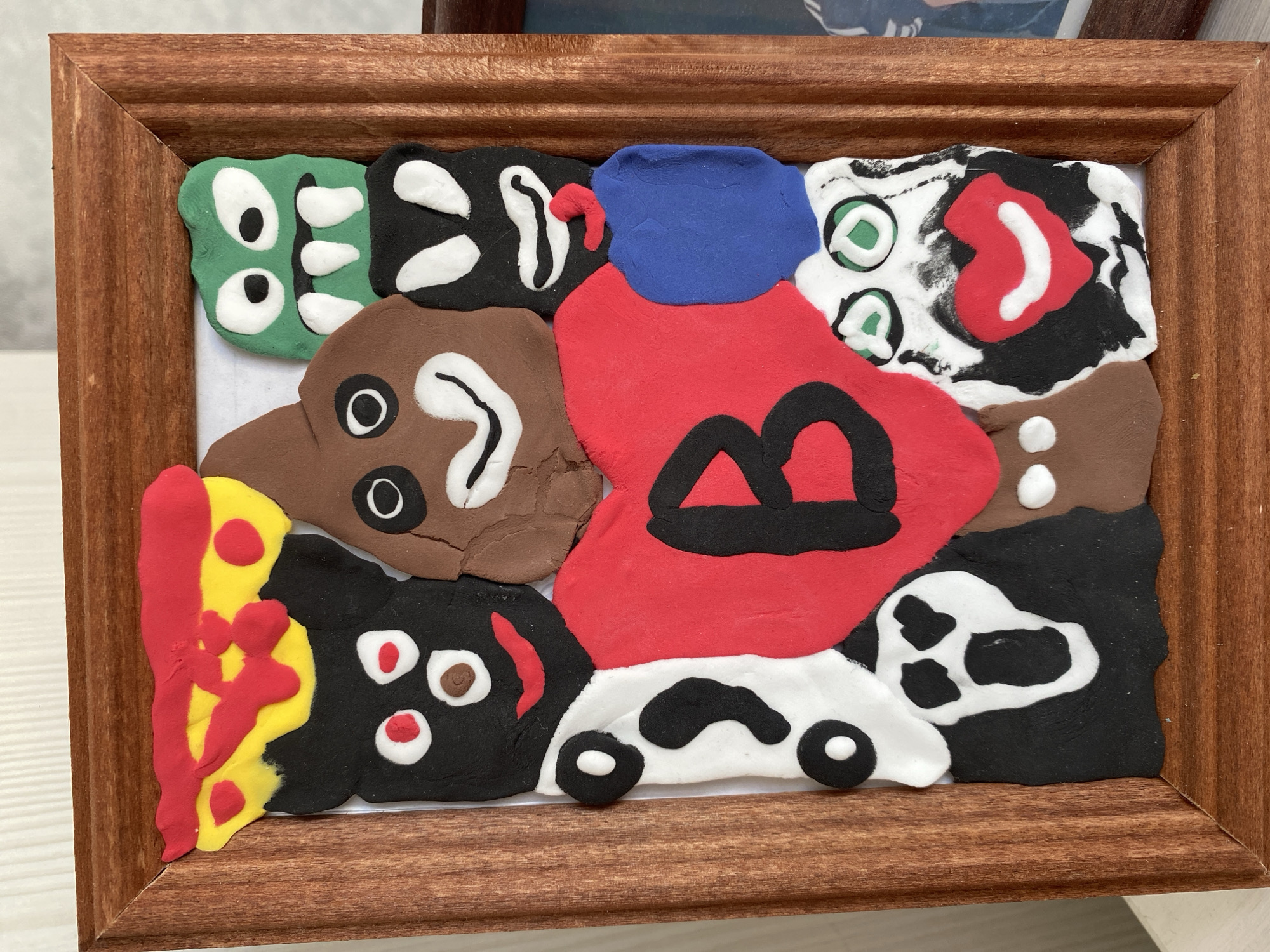 The image displays a framed piece of artwork made from colorful modeling clay. The artwork is a collage of various clay figures and shapes arranged on a white background. In the center is a large red heart with the black letter "B" inscribed on it. Surrounding the heart are several distinct figures: a green monster-like creature with large eyes and sharp teeth at the top right, a brown poop emoji-like figure with eyes and a smiling mouth at the top center, a black figure with white eyes and a red smile at the top left, another black and white figure resembling a ghost or a skull with a gaping mouth at the bottom left, a black figure with white eyes and a mouth below the green monster, and a figure with a white face, green eyes, and a red smiling mouth with a black beard at the bottom right. There are also smaller brown and blue clay shapes. The artwork is housed in a brown wooden frame. The background visible outside the frame is a white surface, suggesting the artwork is placed on a table or shelf. No specific location in the city of Uchaly, Russia, is depicted or indicated in the image.
