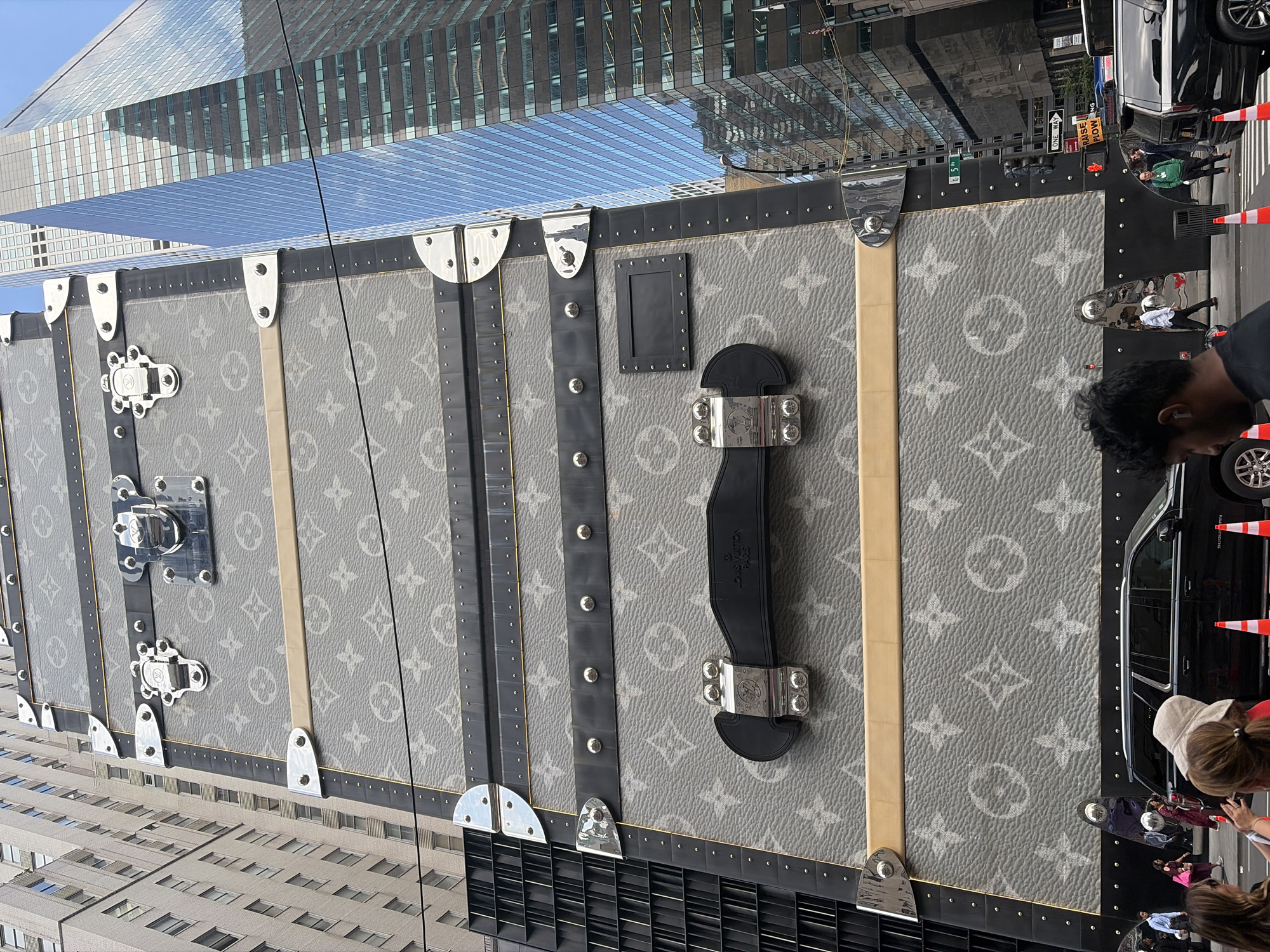 This media file shows a large, grey Louis Vuitton trunk on display in New York City. The trunk features the brand's signature monogram pattern in a lighter grey. It is adorned with silver hardware, including latches and reinforced corners. The trunk is placed outdoors, with a modern skyscraper reflected in its surface and visible in the background. The reflection shows a blue sky and the building's glass facade.
In the foreground and to the right of the trunk, several people are visible. One person is leaning over the trunk, and others are standing around it. They appear to be observing the trunk. The setting suggests a public space, possibly a street or a plaza in a business district given the towering buildings. The lighting indicates it is daytime, likely bright and sunny.
The overall impression is one of an outdoor promotional display or an event showcasing the luxury trunk against the backdrop of the urban environment. The people's casual attire and the presence of traffic cones on the right side of the frame suggest a public street setting. There is text visible on the trunk's black leather strap, which reads "LOUIS VUITTON PARIS".