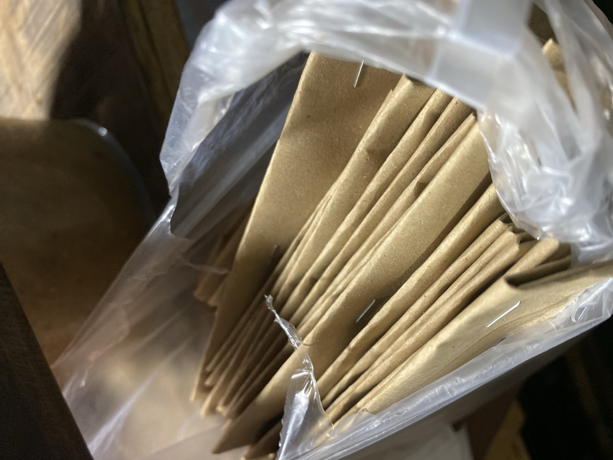 The image shows a close-up, low-angle view of a stack of brown paper envelopes or folders inside a clear plastic bag. The envelopes are neatly arranged, and some have small metal staples visible along their edges. The background is blurred, suggesting a shallow depth of field, but appears to be a wooden surface. There are no people or animals visible in the image, and no specific location in Kasihan, Indonesia, can be identified from the visual information provided. The lighting is dim, casting soft shadows.