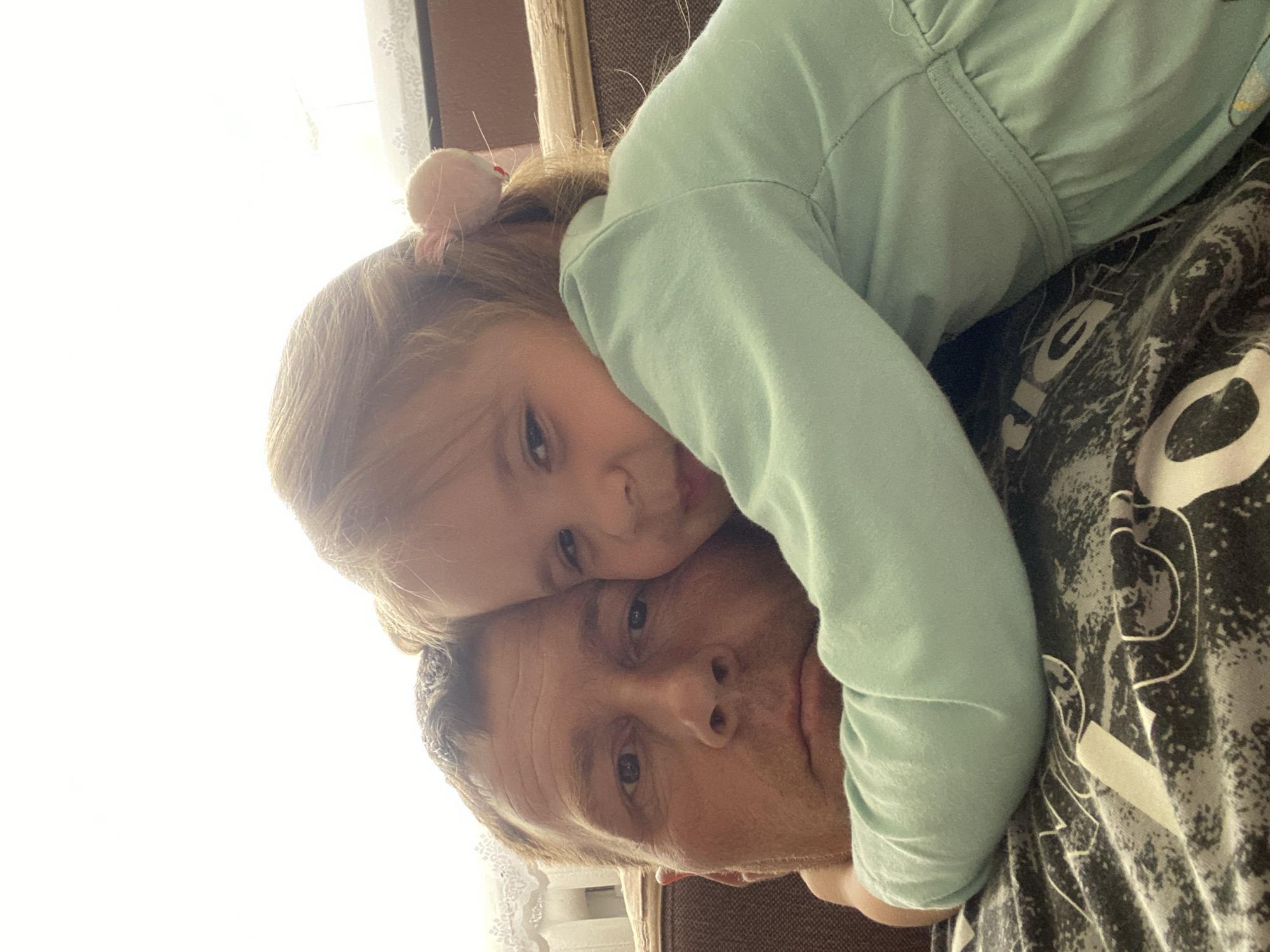 This image, taken in Фоки, Russia, captures a warm moment between an adult man and a young child. The original image is rotated 90 degrees clockwise. When viewed correctly, the man's head and upper torso are visible in the lower right, and the child is leaning across him from the upper left.
The primary subjects are two people. The man, with short hair and some stubble, looks directly at the camera with a calm expression. He is wearing a dark t-shirt with a grey and white abstract, marbling pattern. Partially visible white text on his shirt includes the letters "RIG" and a rounded shape, possibly part of "ORIGINAL" or a similar word. The child, likely a girl, has light brown hair tied into a small bun on top of her head, secured with a light-colored hair tie. She is wearing a light teal or mint green long-sleeved top. Her face is resting close to the man's head, and she also looks towards the camera with a soft, gentle gaze, hinting at a slight smile.
The activity taking place is an affectionate pose, likely a selfie or a close-up snapshot, showcasing a bond between the two individuals. The child appears to be hugging or leaning into the man, suggesting comfort and intimacy.
The setting appears to be indoors, possibly a home, with a bright window area on the left, largely overexposed, indicating strong daytime light. A hint of a white, sheer curtain or lace is visible near the bright light. In the background on the right, blurred details of what appears to be a brown upholstered chair or sofa are visible. The overall mood conveyed by the subjects' expressions is one of contentment and closeness. There are no clear weather indicators beyond the bright indoor light.