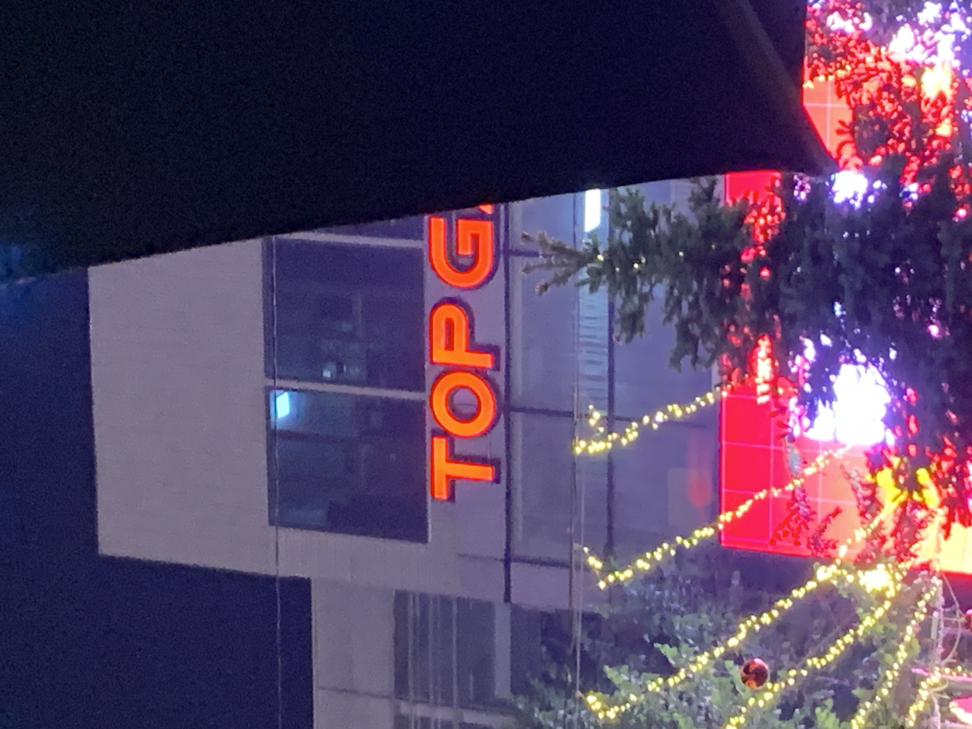 The image shows a nighttime view of a building with "TOP G" lit up in orange letters on its side. A decorated Christmas tree with white and red lights partially obscures the view. The location is likely in the city of Almaty, Kazakhstan, though this cannot be determined from the visual data in the image alone.