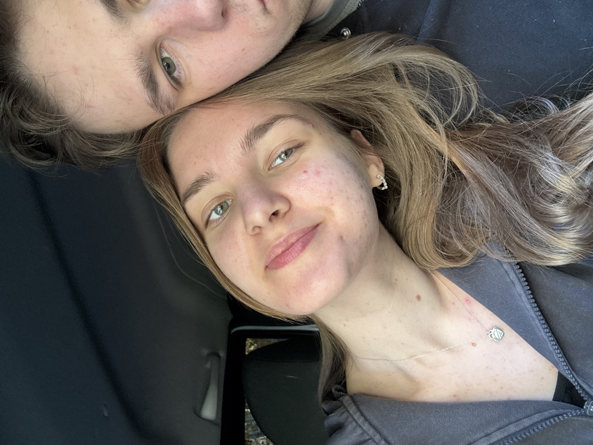 A selfie-style photo shows two individuals, a male and a female, seemingly lying down. The male is at the top of the frame, his forehead close to the female's hairline. He has light brown hair, fair skin, and green eyes. The female is slightly angled towards the camera. She has long, wavy blonde hair, fair skin with some visible blemishes, and green eyes. She is wearing a silver necklace with a pendant and small hoop earrings. They appear to be inside a vehicle, as suggested by the car seat and interior visible in the background. The overall lighting is natural and soft. It is not possible to ascertain their location as the city of Perm, Russia, based on this image alone.