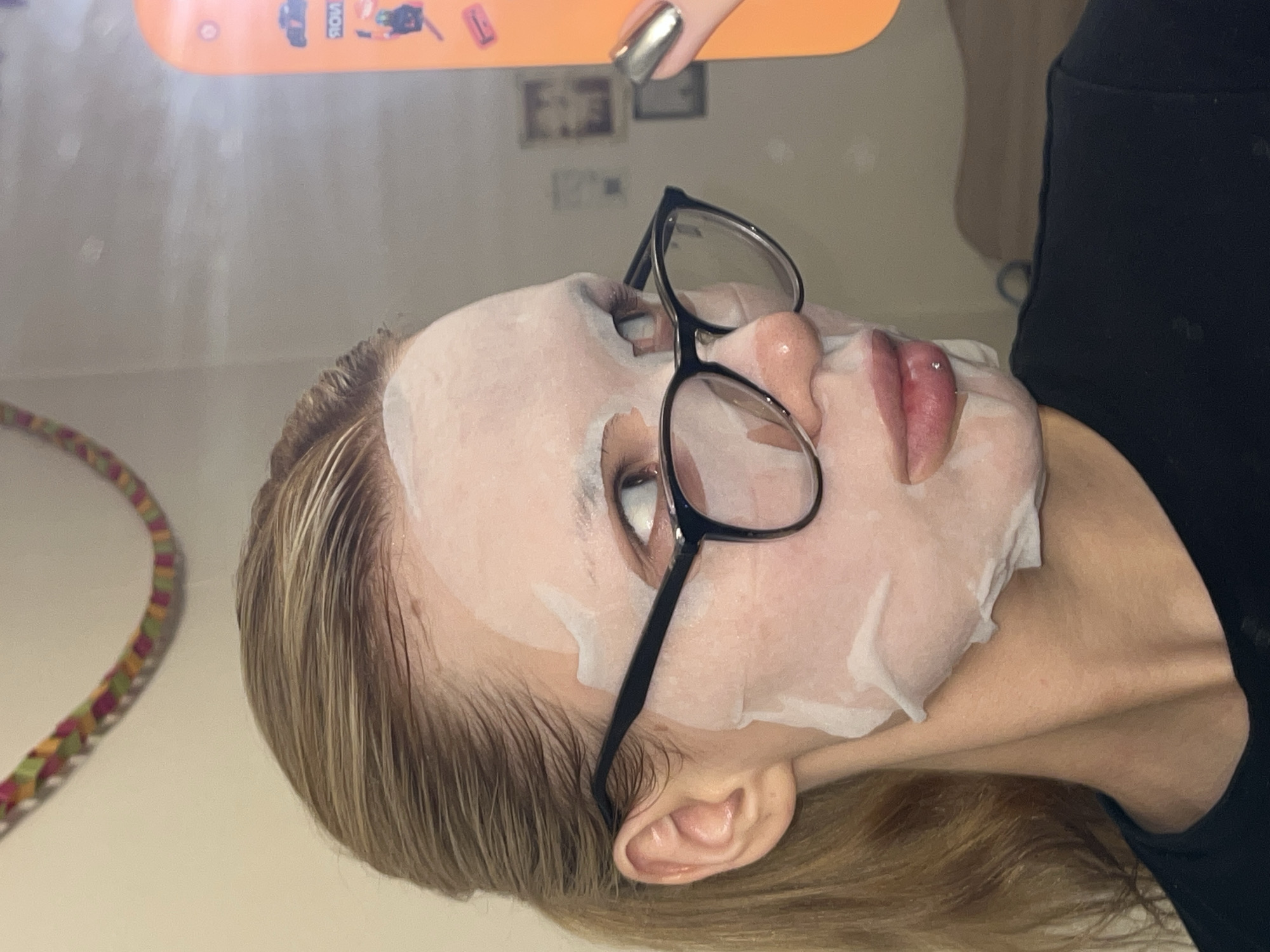 A woman with blond hair is wearing a white sheet face mask and black-framed glasses. She is holding an orange phone in her right hand, with a silver fingernail visible. The background includes a wall with framed pictures and a possible cord. The location cannot be determined from the image.