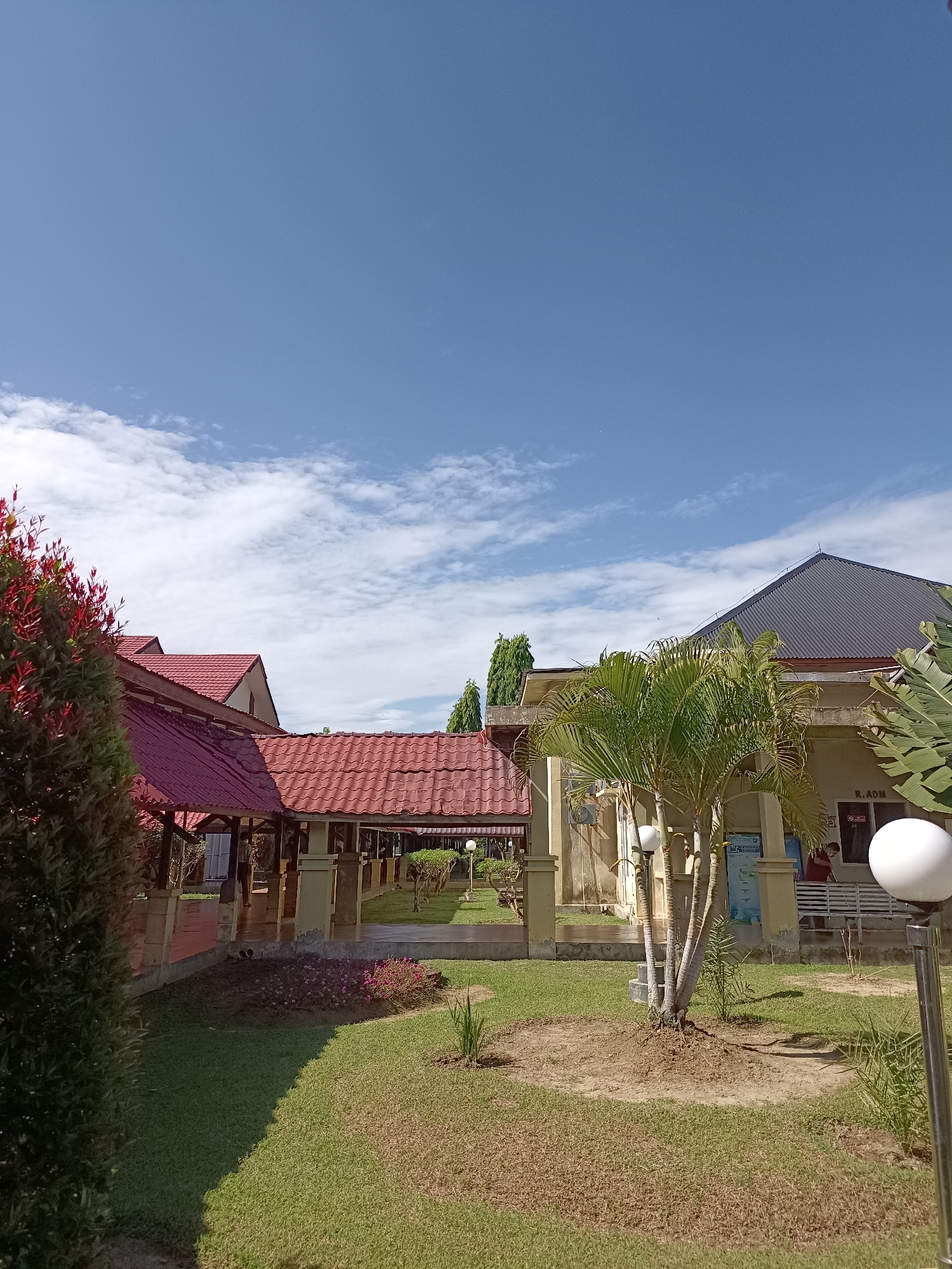 The image depicts an outdoor scene with a clear blue sky dotted with scattered white clouds. In the foreground, to the left, is a dark green shrub with red-tipped foliage. Adjacent to this shrub is a building with a red tiled roof and covered outdoor seating areas supported by pillars. Further to the right, another building features a dark grey metal roof and a light beige facade. A small palm tree stands in front of this building, and a white spherical lamp is visible on a pole in the right foreground. A sign reading "R.ADM" is affixed to the beige building. A white bench is positioned near the beige building, with a person wearing a red shirt and a mask visible sitting on it. The ground is covered with grass, some of which appears dry and brown, interspersed with patches of greener grass and some low-lying pink flowers. The overall setting suggests a public space, possibly a campus or administrative area, in Mesjid Reuntoh, Indonesia.