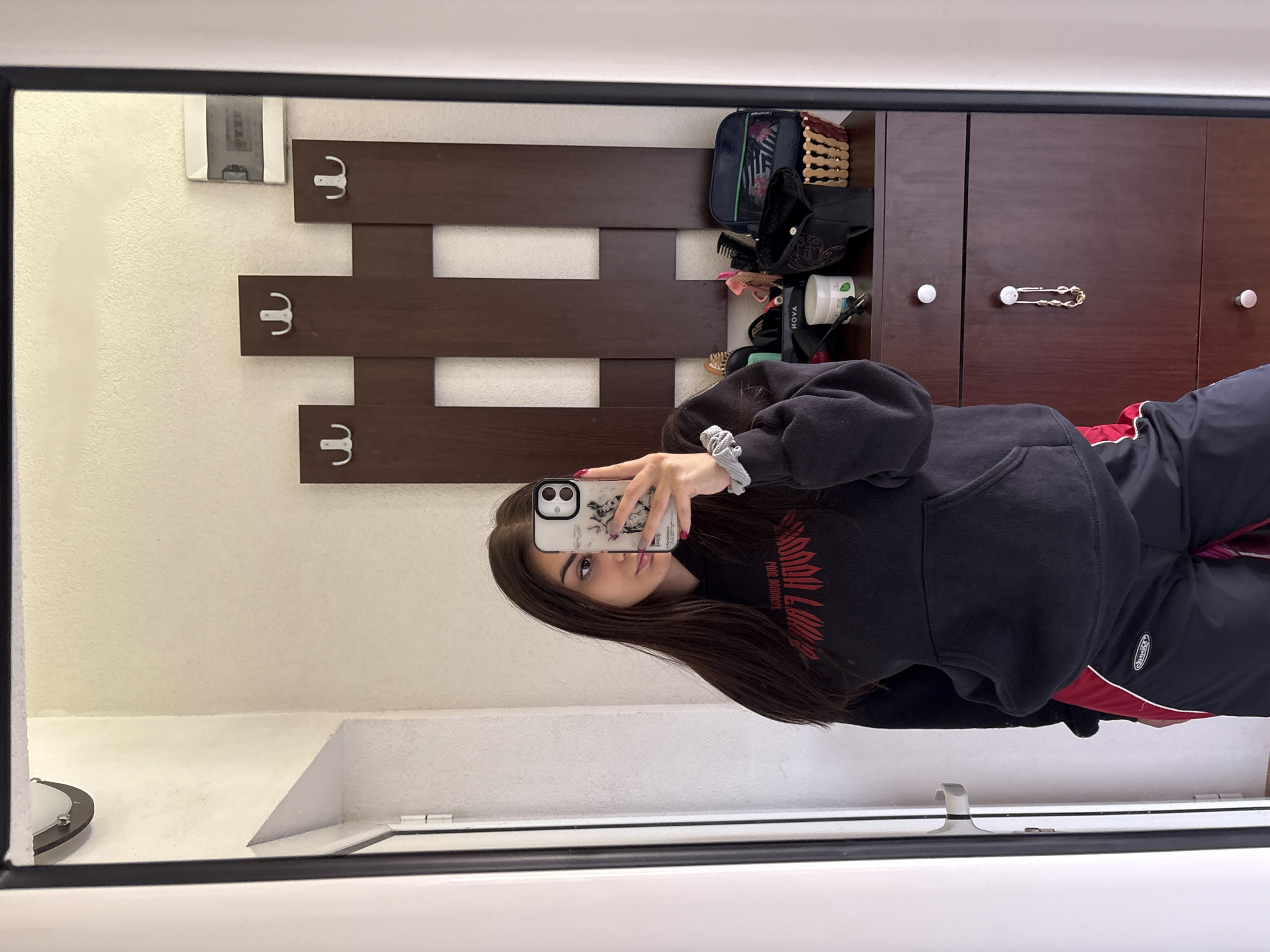A medium shot depicts a young woman taking a selfie in a mirror. The reflection shows part of a room with a white wall and a dark brown wooden coat rack with hooks. Above the coat rack is a dark brown cabinet with white knobs and a decorative safety pin with beads. On top of the cabinet, there's a cluttered arrangement of items, including a bag, hairbrushes, and various containers. The woman is wearing a black hoodie with a red design and dark-colored sweatpants with a white stripe and red trim. The mirror frame is black. It is unknown if the location is in Polena, Bulgaria.