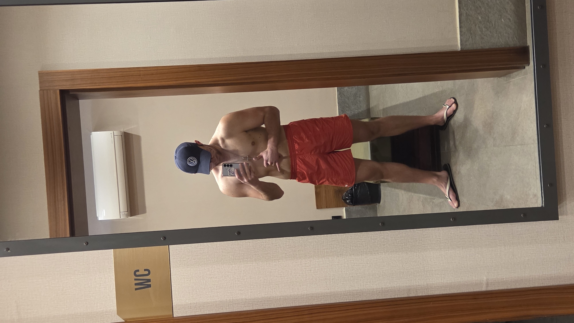 A shirtless man wearing a blue baseball cap and red swim trunks is standing in front of a mirror taking a selfie with a smartphone. The man is also wearing flip-flops. The mirror is in a room with beige walls, a wooden frame, and a metal border. On the wall to the left of the mirror, there is an air conditioning unit and a sign that reads "WC". A black bag is visible on the floor in the reflection of the mirror. The location is Unknown, Unknown.
