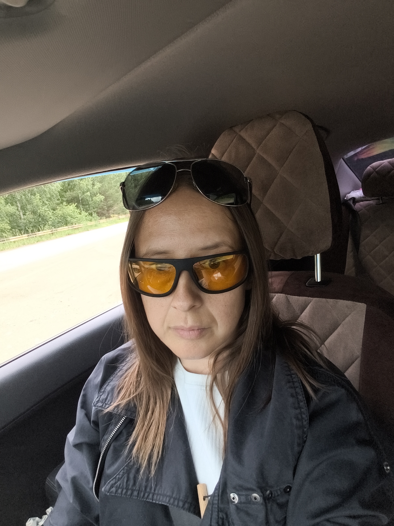 A woman with brown hair, wearing a dark jacket and a light blue t-shirt, sits in the passenger seat of a car. She has sunglasses on her face and another pair of sunglasses on top of her head. The car's interior is visible, with quilted headrests. Outside the car window, a dirt road and trees can be seen, suggesting a rural or suburban setting, potentially near Dubovaya, Russia. The image appears to be a selfie taken from the passenger seat.