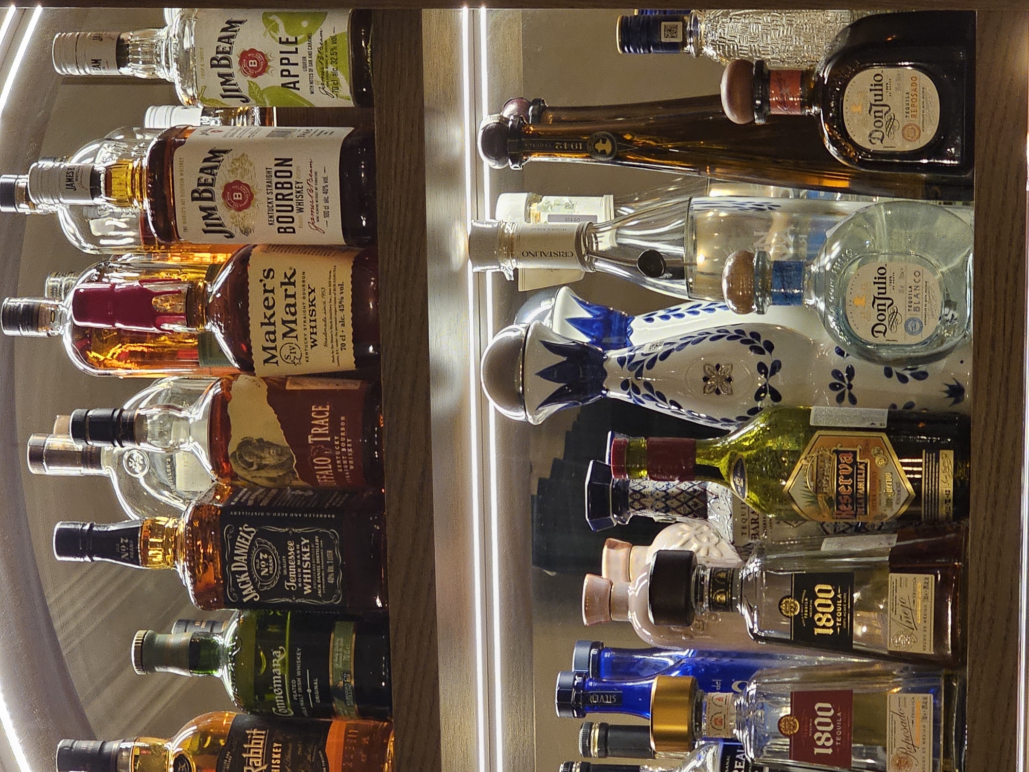 The image shows two shelves of liquor bottles in a bar or restaurant. The bottles on the left shelf are primarily whiskey, including Jim Beam, Maker's Mark, Buffalo Trace, and Jack Daniel's. The bottles on the right shelf are primarily tequila, including Don Julio and 1800. The bottles are stacked, and the shelves are illuminated by vertical LED strip lights. The exact location in Yerevan, Armenia, cannot be determined from the image alone.