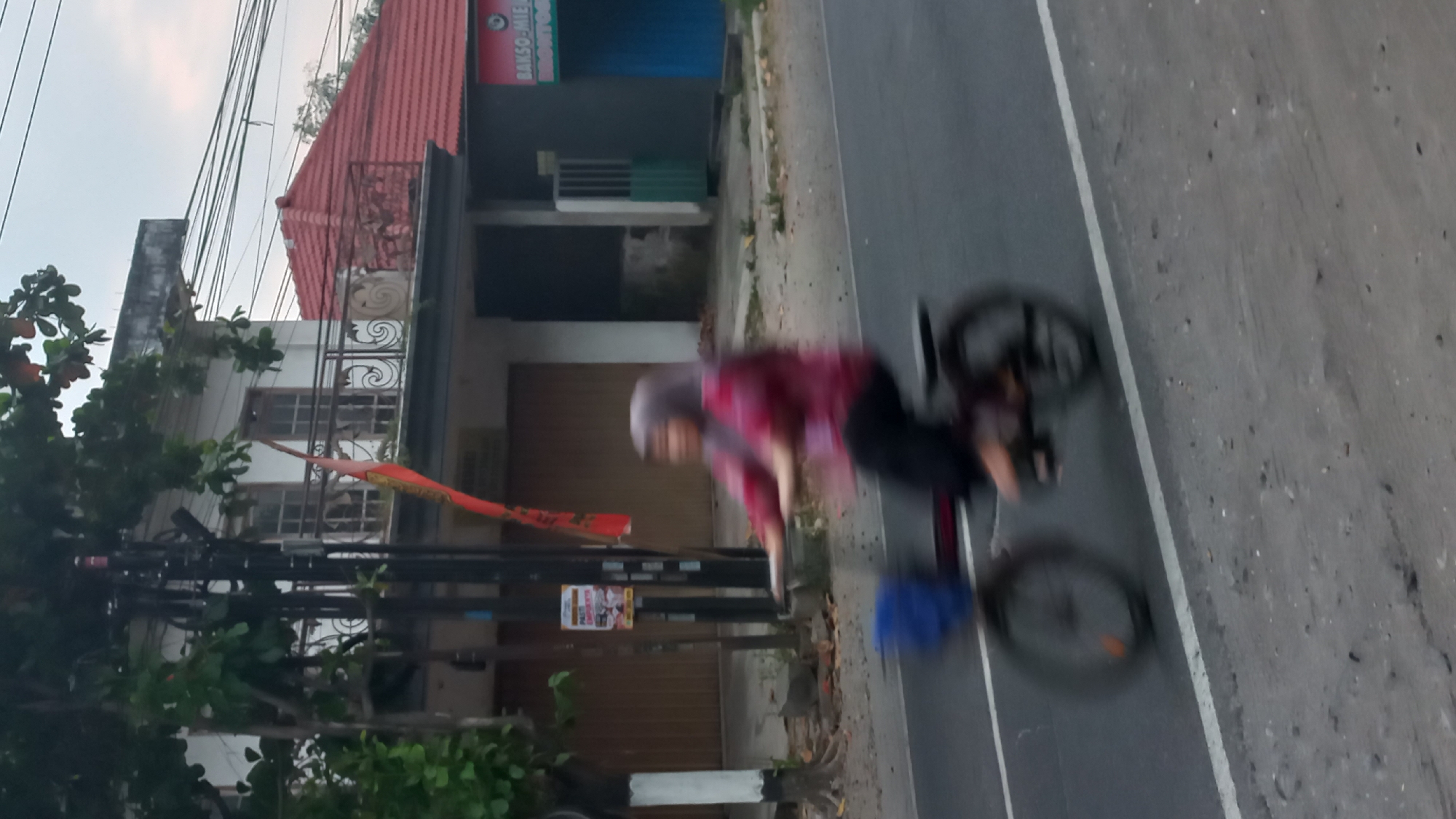 A person wearing a headscarf and a red patterned shirt is riding a bicycle on a paved road in Pare, Indonesia. The person is blurred due to motion, and their face is not clearly visible. The bicycle has a blue basket in the front and appears to be a small, folding model. The road has a white line marking its edge. In the background, there is a building with a white facade and a red tiled roof, with a balcony. There is also a dark-colored building with a sign that reads "BAKSO MIE" and "BRONTO". Trees and power lines are also visible in the scene. The sky is overcast.