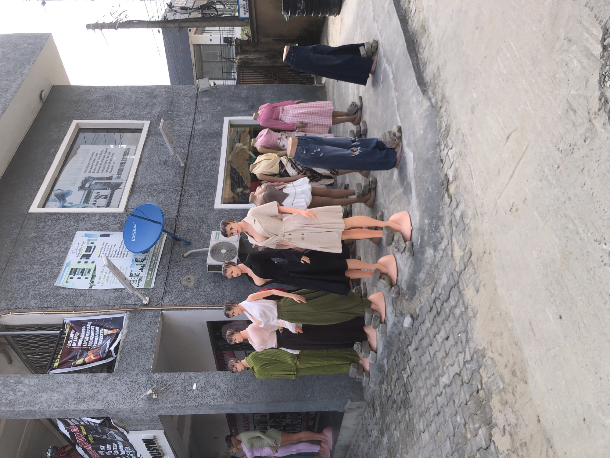 This outdoor daytime scene, likely in a commercial district of Ado, Nigeria, features a line of headless mannequins displaying various clothing items along a paved walkway. The mannequins, some full-bodied and others just torsos or legs, wear a mix of dresses, skirts, pants, and shirts in different colors and styles, standing on small stone blocks.
On the left, a two-story building with a grey, textured facade serves as a backdrop. Its ground floor features several open windows or doorways. One banner advertises "SALES OF +DELL LAPTOP, HP LAPTOP, MAC & LENOVO LAPTOP BATTERIES, ACCESSORIES, POWER BANK, INVERTER." Another smaller sign indicates "boutique," and text "CYP CAD DESIGN USTIN" is also visible.
The first floor of the building has a large window displaying services like "Digital Coloured Prints, cards, Brochure, Graphic Design, Binding, Business Cards, Photocopy, Computer Training, Cyber Caf, Lamination, Gift items, Phone" with a contact number "Tel: 08:308107880782670556". Mounted on the building wall are a blue DStv satellite dish and an outdoor air conditioning unit. Another poster on the wall promotes "Randburg Estates," announcing "NOW SELLING 2 BEDROOM LUXURY APARTMENTS" with amenities like "Swimming Pool, 24/7 Electricity, 24/7 Security, Smart Home, Electric Fence, Elevator, Installed Inverter, Undercover Park," and providing a contact number "CONTACT: 08167424512, 0783454234)".
The ground surface transitions from grey interlocked paving blocks to a lighter, possibly concrete or sandy area. The sky appears bright, suggesting a sunny day. A utility pole with wires is visible in the upper background.