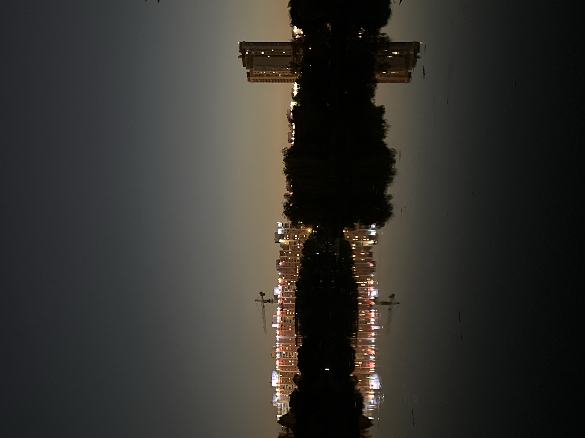 The image depicts a nighttime cityscape scene with a body of water in the foreground. The sky is a gradient of dark gray to lighter tones near the horizon. A line of buildings with illuminated windows is visible across the middle ground, with a construction crane positioned among them. To the right, a taller, distinct building with visible lights stands out. The dark silhouettes of trees and foliage are present between the water and the buildings. The surface of the water reflects the lights from the buildings, creating a distorted but recognizable mirrored image. The overall impression is one of urban development reflected in a tranquil, dark body of water at dusk or dawn.