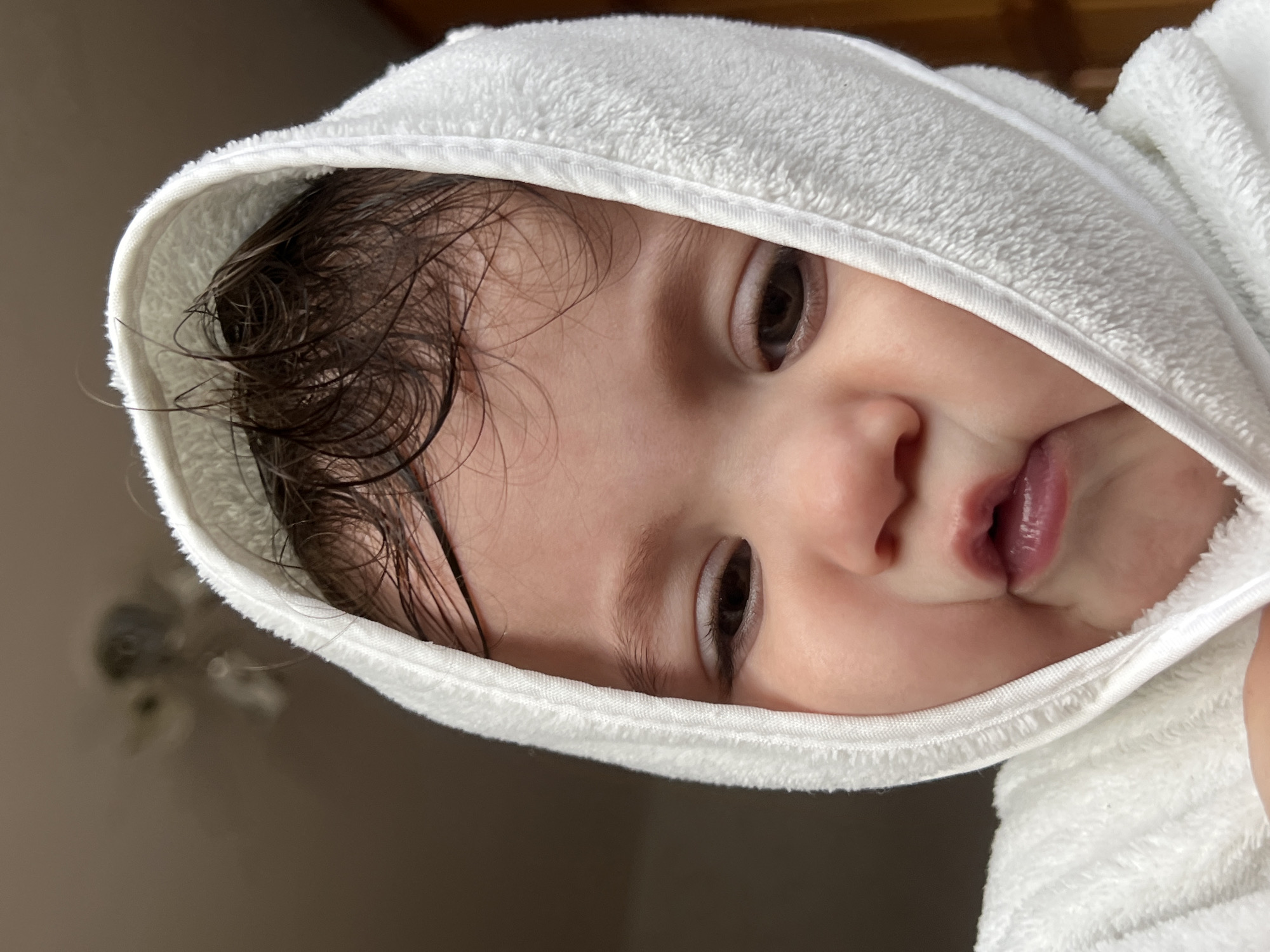 A close-up, high-angle portrait shows a baby or very young child wrapped in a white, hooded towel, likely after a bath. The image is oriented so the child's head is significantly tilted within the frame. The child's dark, wet, and slightly curly hair emerges from under the towel. Their large, dark brown eyes are open wide, looking directly at the camera with a neutral, perhaps curious, expression. The child's small nose is visible, and their lips are slightly parted. The white towel is made of soft terrycloth, framing the child's face and contrasting with their fair skin. The background is a blurred, plain light brown or beige indoor surface. No specific location cues are visible within the image, beyond the implied indoor setting. There is no visible text. The photograph was taken in Elena, Bulgaria.