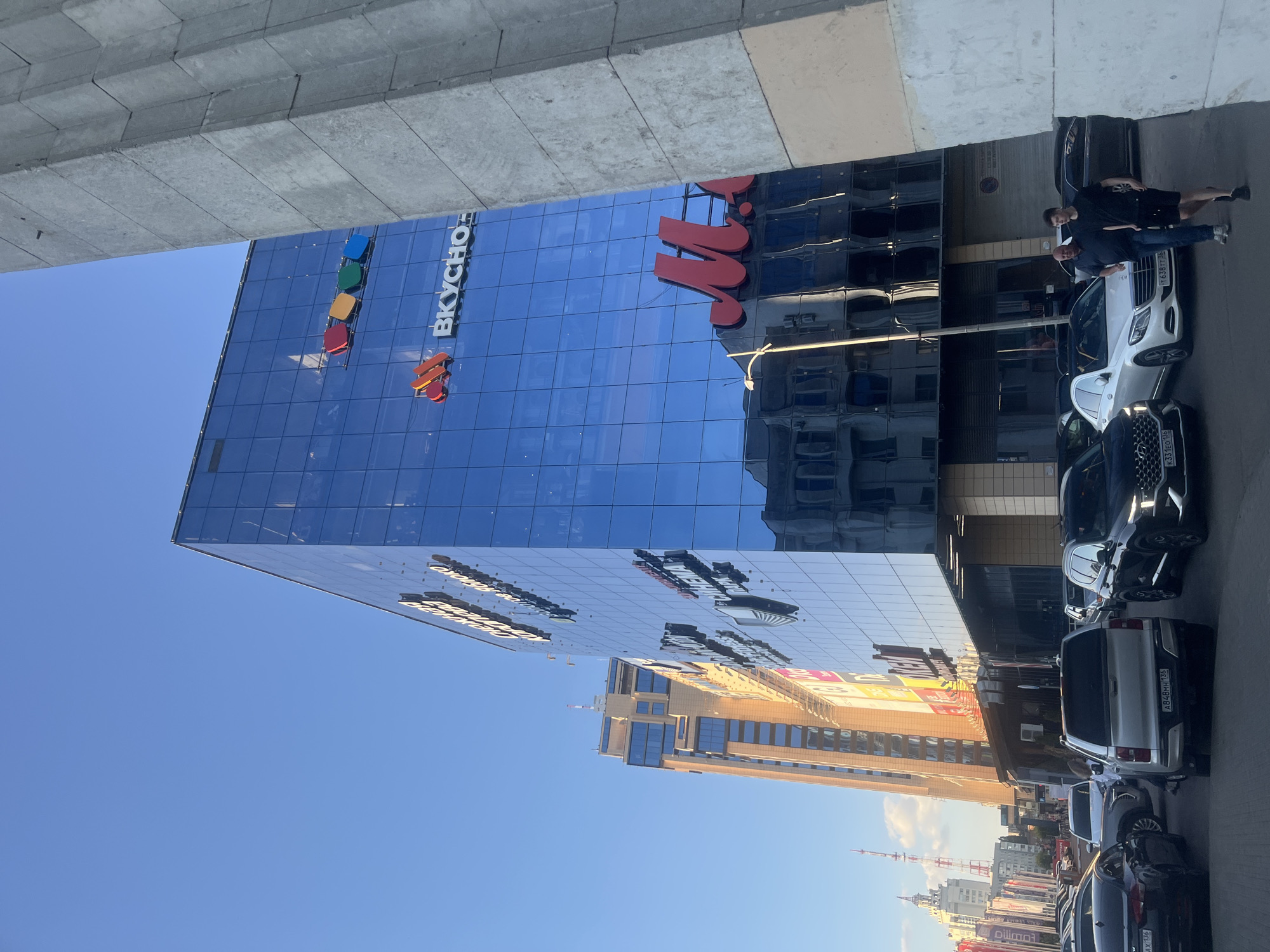 The image depicts an exterior scene featuring a modern glass-fronted building, likely a commercial or office space, with a clear blue sky overhead. The building's facade is highly reflective, mirroring the surrounding environment, including other buildings and the sky. A prominent sign on the building reads "ВКУСНО" (Vkusno), which translates to "tasty" or "delicious," suggesting it might be related to a food establishment or a brand. There are also colorful square logos on the building.
In the foreground, several parked cars are visible, lined up along the street or a parking area. Two individuals are standing near a white car, one in a dark t-shirt and shorts, and the other in a dark shirt and jeans. Their postures suggest they are conversing or observing something. The time of day appears to be daytime, judging by the bright sunlight and the clear sky, possibly late afternoon or early evening due to the angle of the shadows. The setting is urban, indicated by the presence of the buildings and cars.
The overall impression is one of a contemporary urban environment under pleasant weather conditions. The reflections on the building's glass add a dynamic element to the scene. The presence of the sign "ВКУСНО" provides a specific clue about the nature of the establishment or business housed within. The location context provided, Новоподклетное, Russia, further grounds the scene.