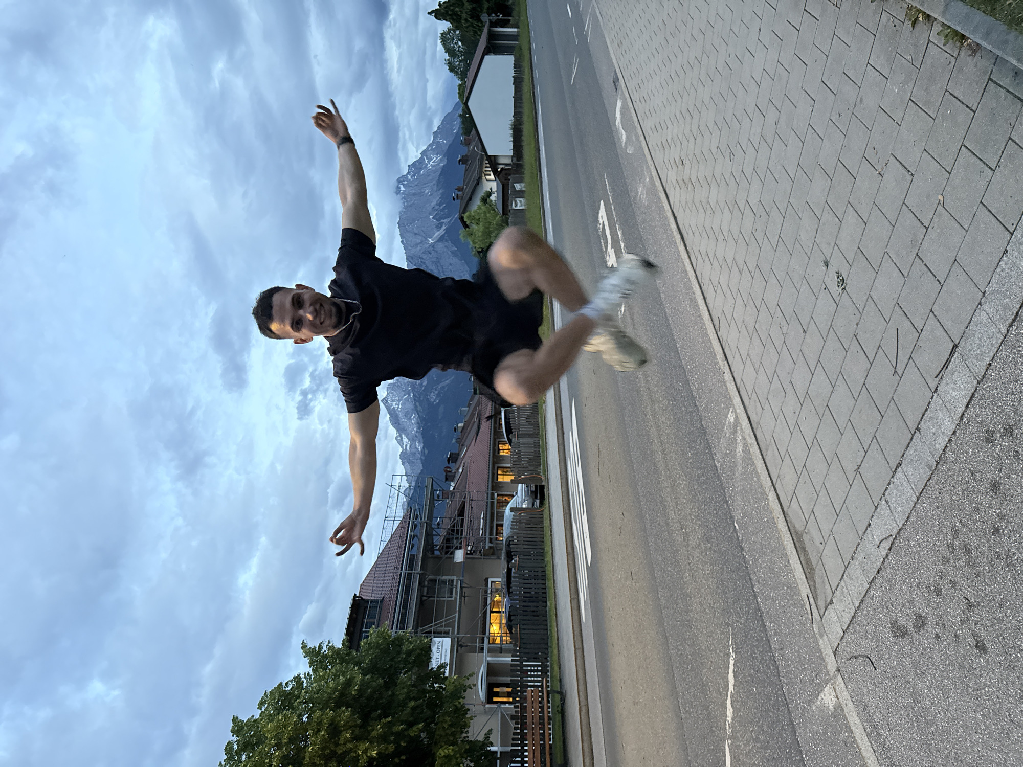 A young adult male, dressed in a black t-shirt, black shorts, and light-colored athletic shoes, is captured mid-jump above a paved road. His arms are extended horizontally with open hands, and his legs are bent, bringing his feet up behind him. A silver-colored chain necklace and a dark wristband on his left wrist are visible. The scene is located in Garmisch-Partenkirchen, Germany, evidenced by the prominent, rugged, snow-capped mountains in the distant background, characteristic of the Wetterstein Mountains, including the Zugspitze massif.
The immediate background features a two-lane paved road with a white-striped pedestrian crosswalk. To the left, a multi-story building with a red tiled roof is partially obscured by a large green tree. Scaffolding is erected on the building's facade, indicating ongoing work, and several windows on the ground floor are illuminated. A wooden bench is positioned in front of a wooden fence next to the road. To the right of the road, a grey paved sidewalk with rectangular pavers is visible. Beyond the road and closer to the mountains, several residential structures with red roofs are present. The sky is overcast with dense grey clouds, suggesting either dusk or an overall cloudy day. The ambient light is diffused.