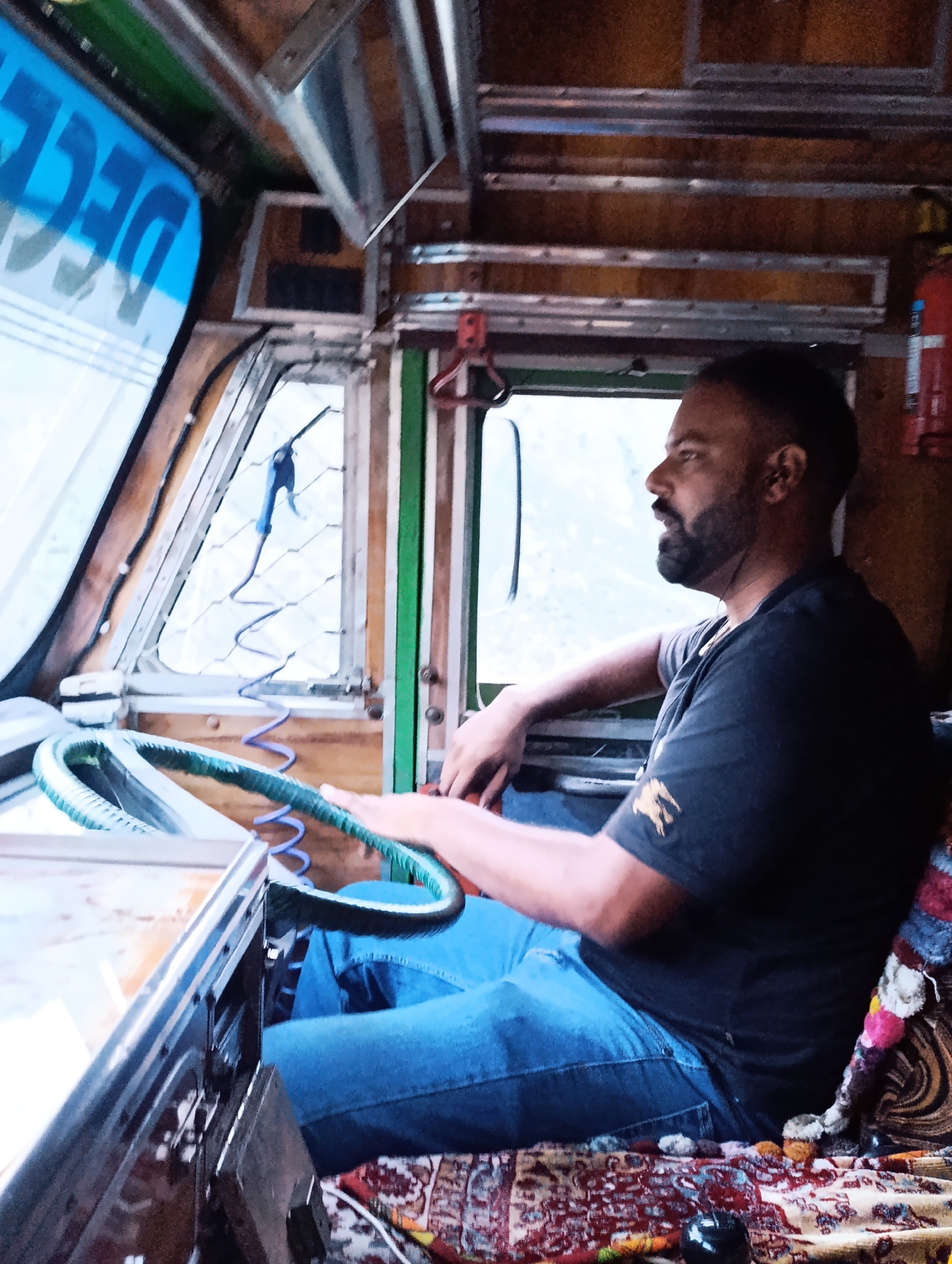 A man is sitting in the driver's seat of a truck. The interior of the truck is visible, with wooden paneling and metal shelving. The man is wearing a dark t-shirt and blue jeans. He has a beard and is looking out the window. A steering wheel is in front of him, and a fire extinguisher is mounted on the wall. The windshield has blue lettering visible on it. The man appears to be in motion, as suggested by the blur of the background outside the window. The scene is likely a roadside stop or a parking area in Kead, India, given the context of the image.