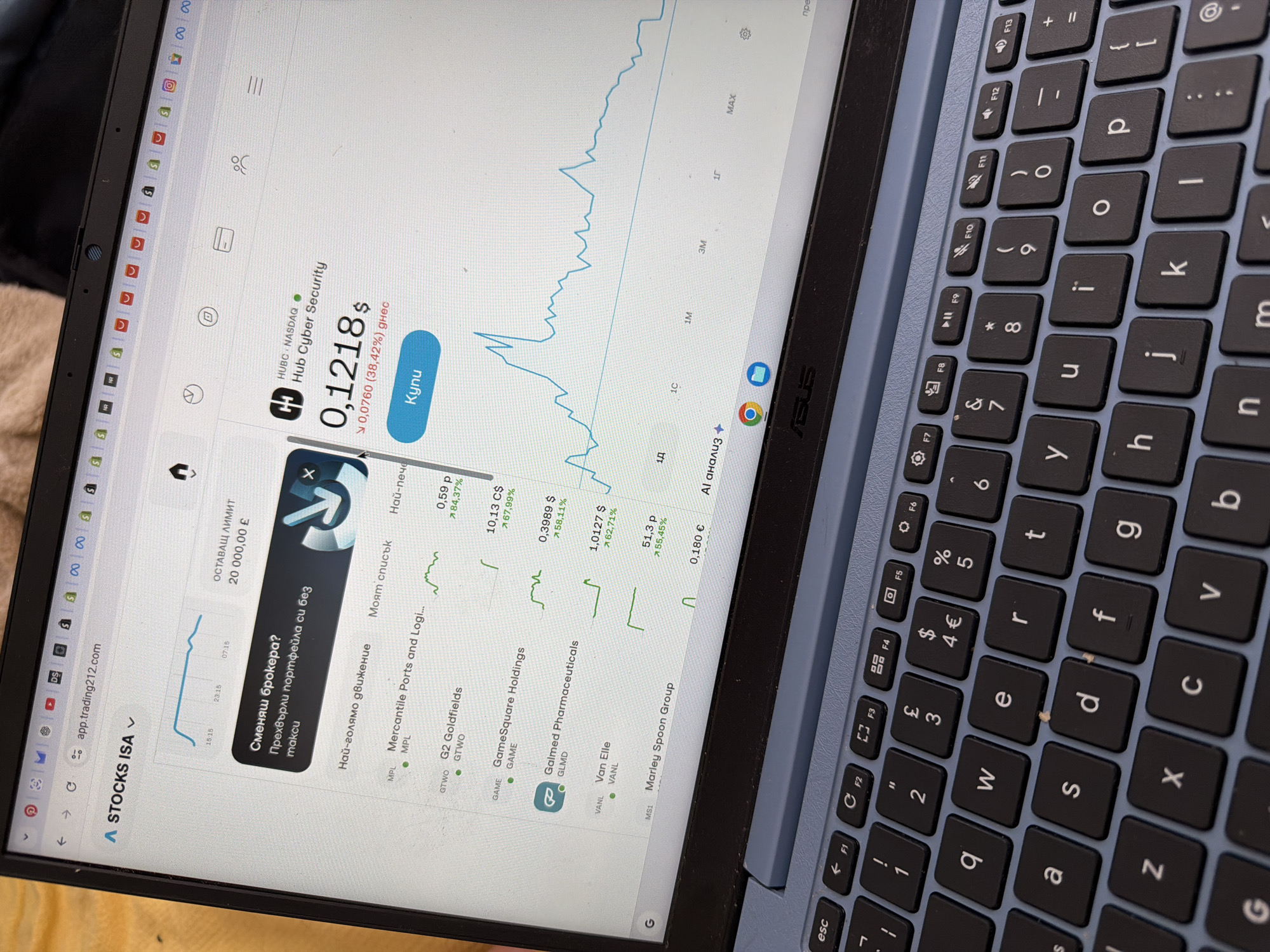 The image shows a laptop screen displaying a stock trading platform. The website, "app.trading212.com", is open on a stock trading application showing stock prices and stock market charts. Specifically, "Hub Cyber Security" is highlighted, its current value at $0.1218.
The laptop is an ASUS, with a black keyboard with blue keys and a silver body. The photo is taken indoors.
I am unable to locate the precise geolocation within Pentre, United Kingdom from this image.