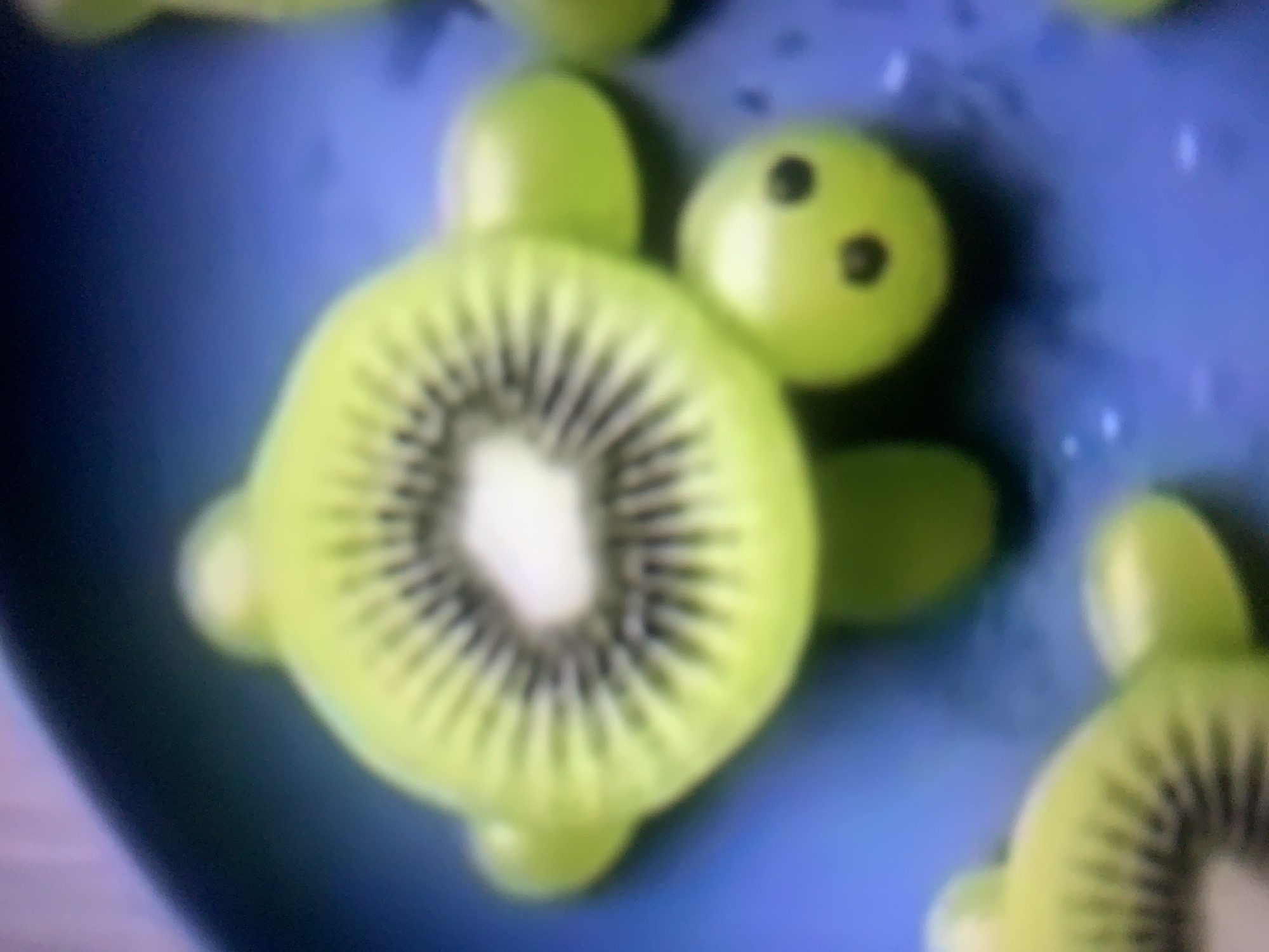 A close-up, blurred image shows a food art creation resembling a turtle on a blue plate. The turtle's body is made from a slice of kiwi fruit, showing its characteristic seeds. Four green grapes are used as the turtle's legs and head. Two of the grapes, forming the head, have two dark spots each, representing eyes. The background is out of focus, suggesting a shallow depth of field. The image does not provide any information about the city of Almaty, Kazakhstan.