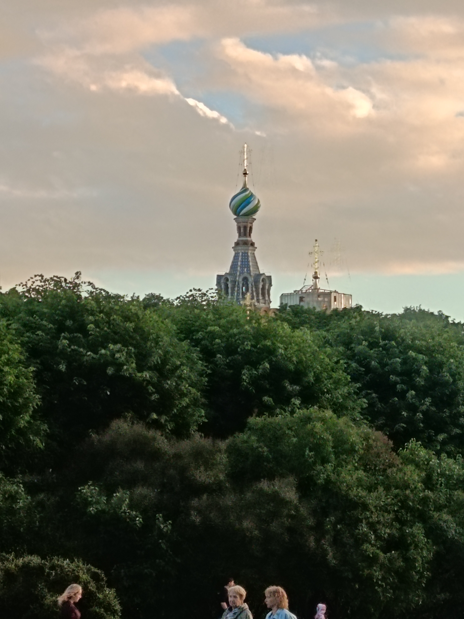 The image shows a view of the St. Andrew's Cathedral in Saint Petersburg, Russia. The cathedral's distinct blue and green domed spire is visible above a dense canopy of green trees. The sky is a soft, hazy mix of pink and blue clouds. In the foreground, several people are casually walking and standing amidst the foliage. The overall impression is one of a peaceful, late afternoon or early evening scene in a park or garden setting with the iconic cathedral spire serving as a prominent landmark in the background.