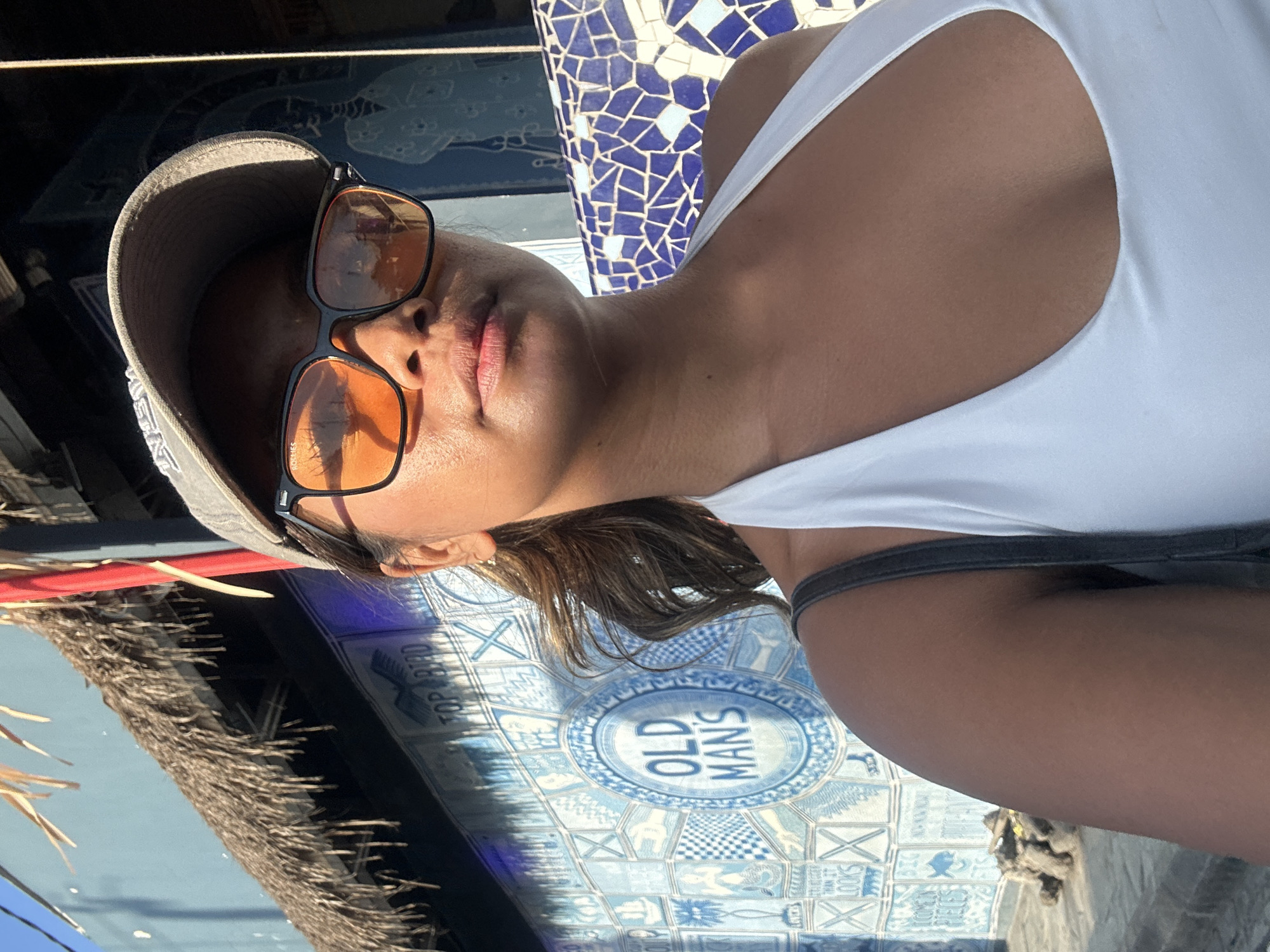A young woman with dark hair pulled back in a ponytail, wearing an orange-tinted pair of sunglasses and a grey baseball cap, takes a selfie in front of a blue and white tiled wall in North Kuta, Indonesia. The wall features a circular emblem that reads "OLD MAN'S". She is wearing a white halter top. To the left of the woman, a thatched roof overhang is visible. The lighting suggests it is daytime.