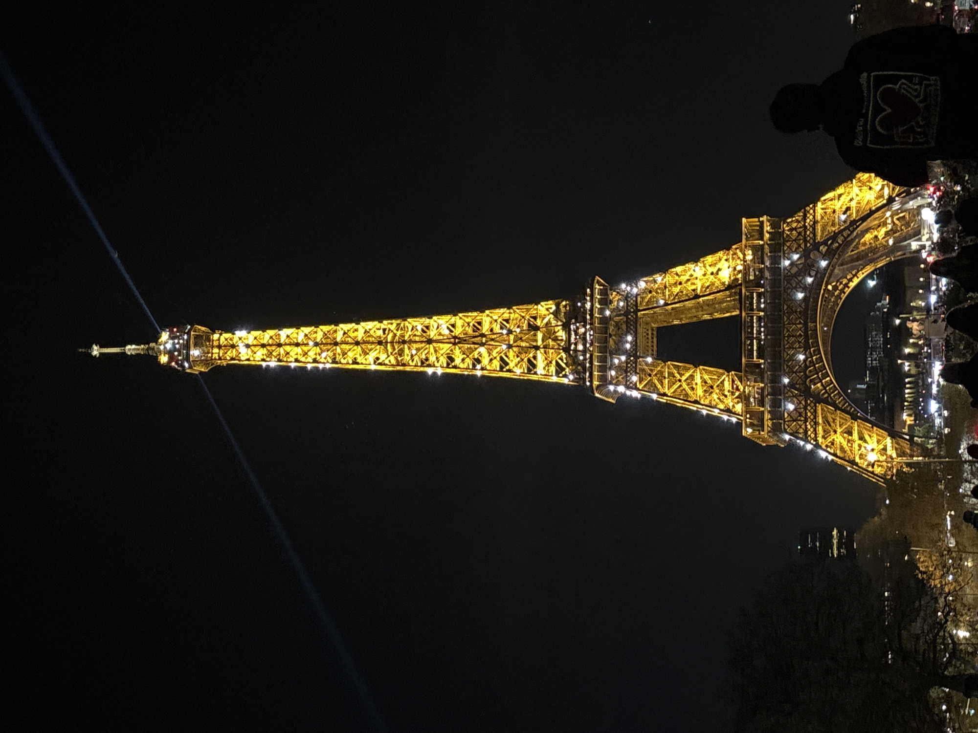 The Eiffel Tower is illuminated at night in Paris, France. A bright spotlight beam shines from the top of the tower into the night sky. The tower's structure is accented with golden lighting. To the right, silhouetted figures of people are visible, indicating a viewing area or gathering. Trees and faint cityscape elements are seen near the base of the tower on the right side of the frame.