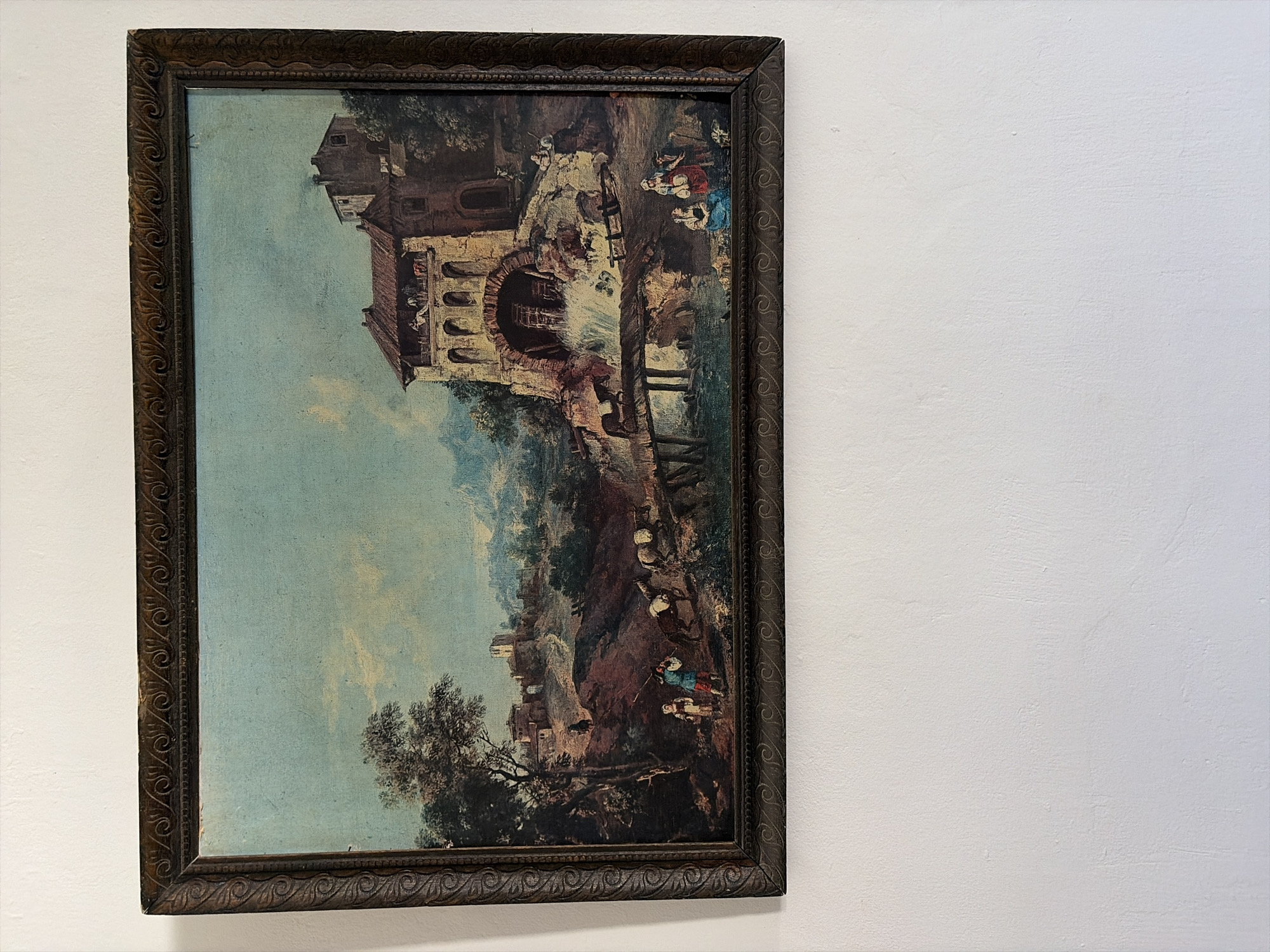 The image shows a framed landscape painting hanging on a white wall. The painting depicts a rural scene with a building in the foreground featuring an archway and multiple stories. People are visible around the building and in the landscape, engaged in various activities. A bridge extends over a body of water, and the landscape includes trees, hills, and buildings in the distance. The painting is within a decorative, dark-colored frame. The location in the city of Unknown, Unknown cannot be determined from the image.