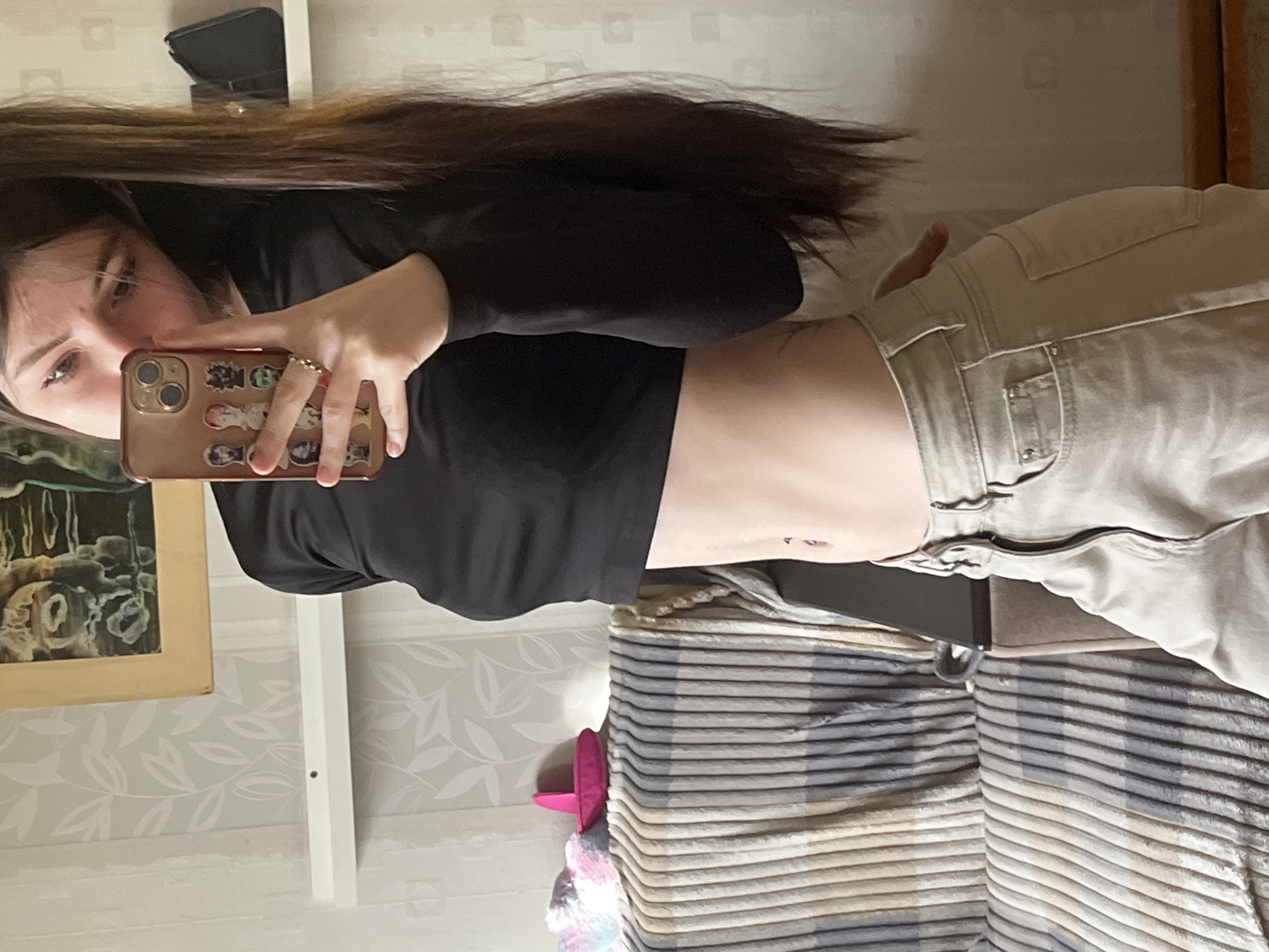 A young woman takes a selfie indoors. She is wearing a black long-sleeve crop top and khaki pants. The phone case is decorated with stickers. She appears to be in a room with patterned wallpaper and a striped bedspread. A painting hangs on the wall. The location is Unknown, Russia.