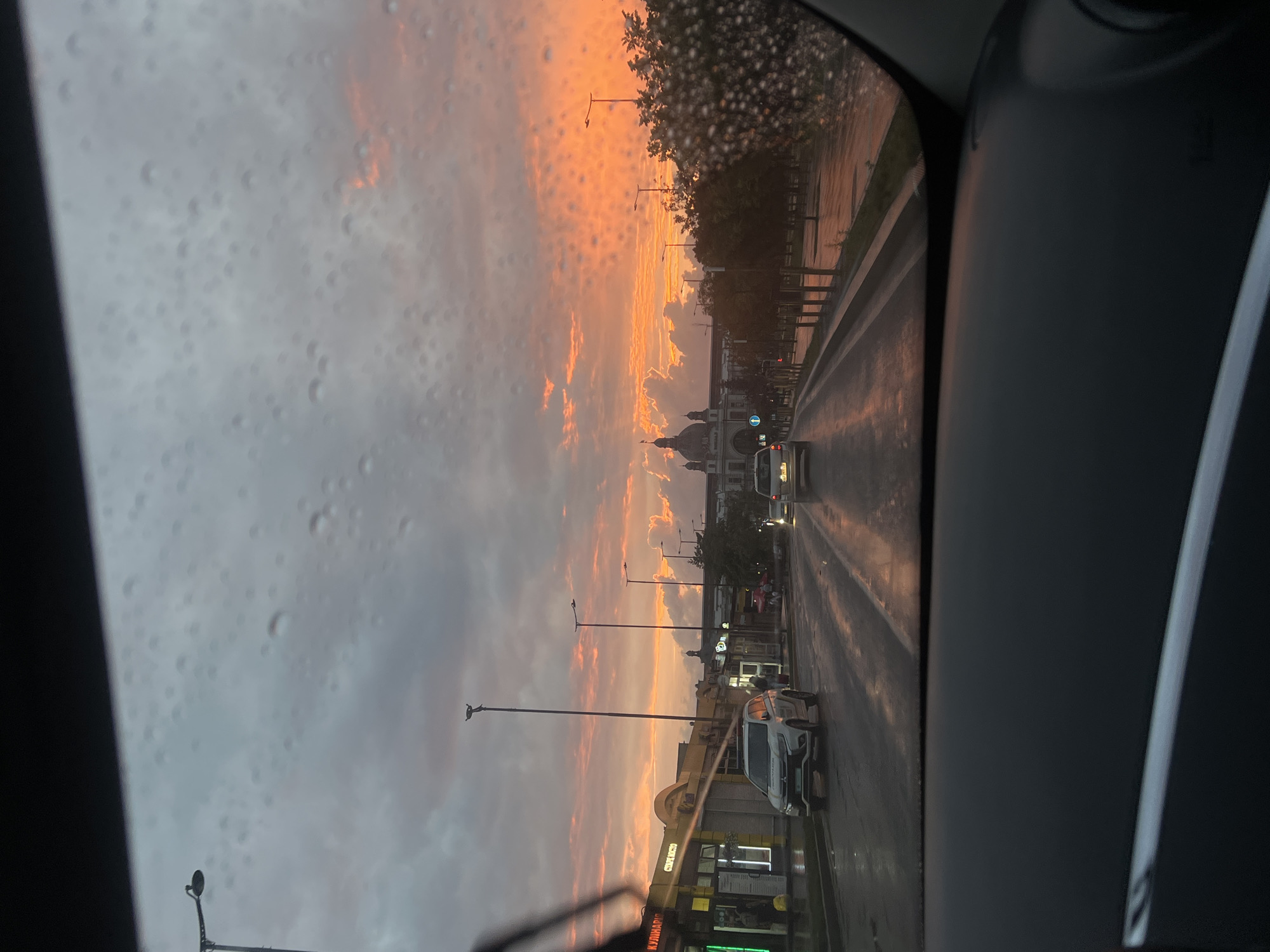 The image depicts a view from inside a vehicle, looking through a rain-streaked windshield at a street scene in Lviv, Ukraine. The sky is ablaze with a vibrant sunset, casting an orange and pink glow on the clouds. The road is wet from recent rain, reflecting the colorful sky and the headlights of the vehicles. On the left side of the street, there are buildings with illuminated signs, including one that says "КУЛІНАРІЯ" (Culinary) and another that says "СТРАХОВЕ МІСТО" (Insurance City). Several cars are driving on the wet road, and streetlights line the avenue. In the distance, a large domed building is visible. The overall impression is of a city street bathed in the dramatic light of a sunset after a rain shower.