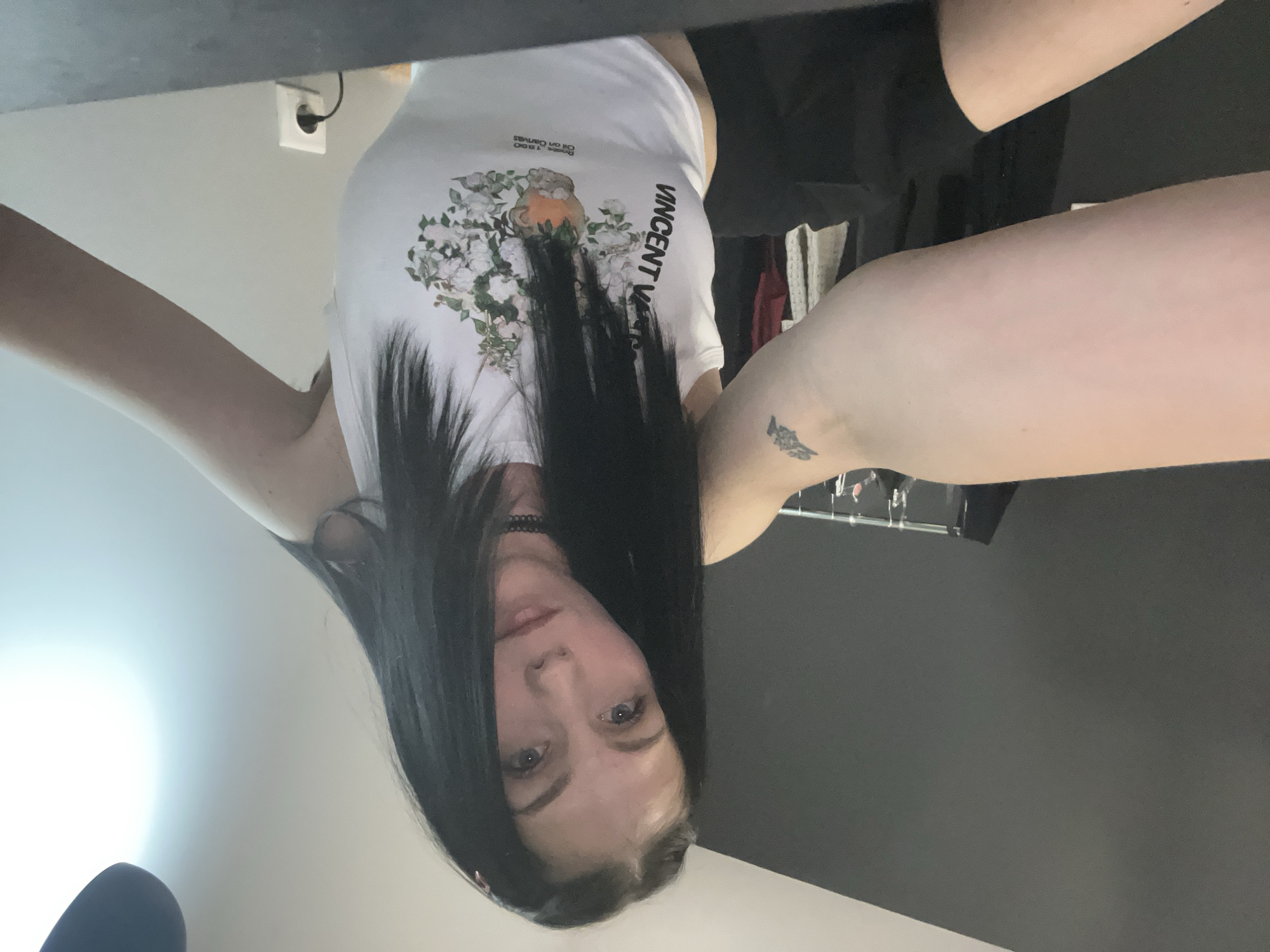 A person, likely female, with long, straight dark hair, is depicted in an indoor setting. She is looking directly at the camera with a neutral expression. The image is presented upside down, suggesting it was taken from a low angle looking upwards, or the photo was taken with the camera inverted. Her arms are raised and spread, possibly holding the camera above her head to take a selfie.
She is wearing a white sleeveless or short-sleeved top and dark shorts. A black choker necklace is visible around her neck. The white top features a graphic print of a bouquet of white flowers and green foliage, with a small, light-colored pot or bird-like figure nestled among the flowers. Text on the shirt includes "VINCENT" written vertically along the right side (viewer's left), and above the floral graphic, the words "Paris 1880" and "Oil on Canvas". A small, dark tattoo is visible on her right upper arm, appearing to be an abstract shape or an animal.
The background is divided into two distinct areas. The upper portion is a light-colored wall or ceiling, where a white wall outlet with a black cord plugged in is visible. The lower portion of the background is a dark grey or black surface or wall. The lighting is bright, creating a strong highlight in the upper left of the image (as presented in the original file). Her eyes are blue or grey.