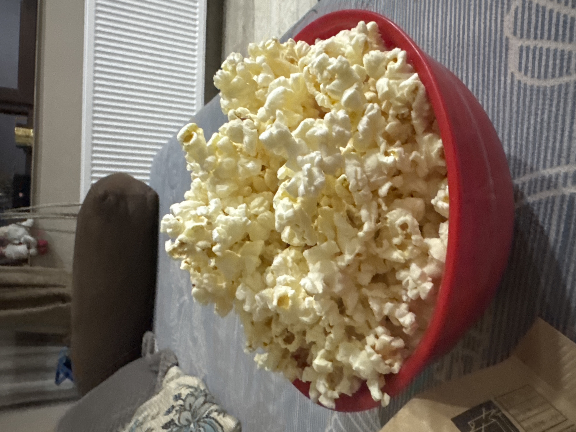 The image shows a red bowl overflowing with popcorn, situated on a light grey couch with a striped pattern. A brown bolster pillow is adjacent to the couch. In the background, there is a window with white blinds. Parts of a shelf with items on it can be seen behind the pillow.