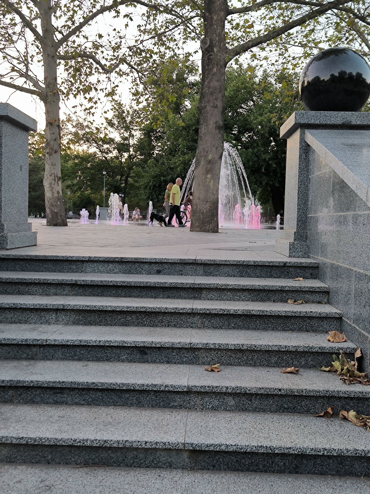 This image depicts a public space with a water feature and people. In the foreground, there are granite steps leading upwards. In the midground, a water fountain with pink-colored water sprays upwards. Two individuals, one in a yellow shirt and another in a darker outfit, are walking near the fountain. A black dog is also present, interacting with the people. Further in the background, more fountains are visible, and trees line the perimeter of the area. The sky appears overcast, suggesting it might be late afternoon or early evening. Based on information associated with the original image, this scene is located in the City of Mykolaiv, Ukraine, likely at a park or public square.