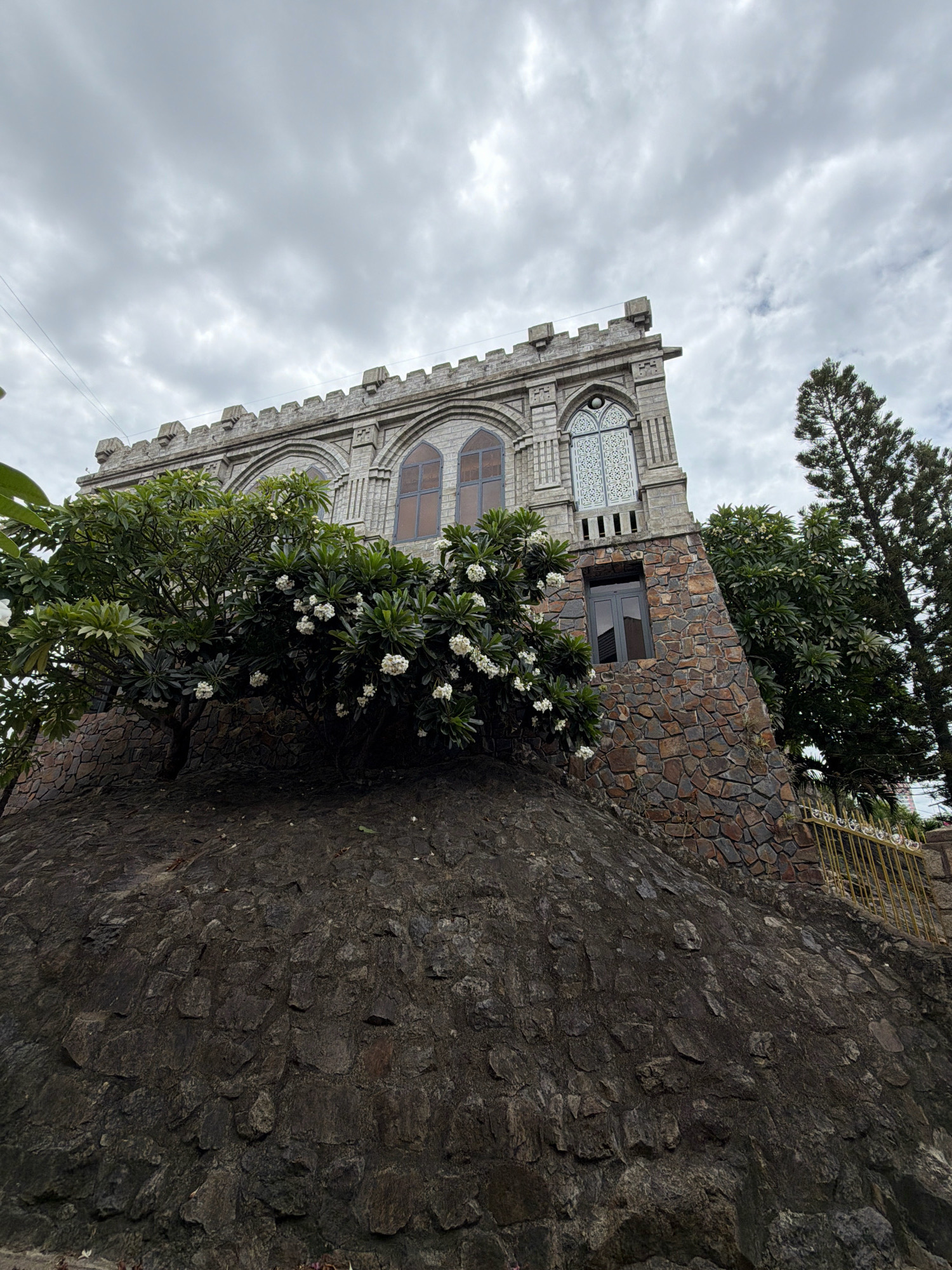 The image depicts a stone building with a castellated roofline, set against a backdrop of a cloudy sky. The building's facade is adorned with arched windows, some featuring intricate decorative patterns, and is constructed from grey stone blocks. The lower portion of the building is covered in rough-hewn stones, creating a rustic appearance. In front of the building, a lush green tree with white flowers is visible, adding a touch of nature to the scene. The building is situated on a rocky incline, with a stone-lined wall and a yellow metal fence visible in the background. This architectural style is reminiscent of a castle or fortress and suggests a historical or decorative purpose. Based on the architecture, particularly the use of stone and the crenellated roof, this building appears to be the "Big Buddha" temple, also known as Long Son Pagoda, located in Nha Trang, Vietnam.