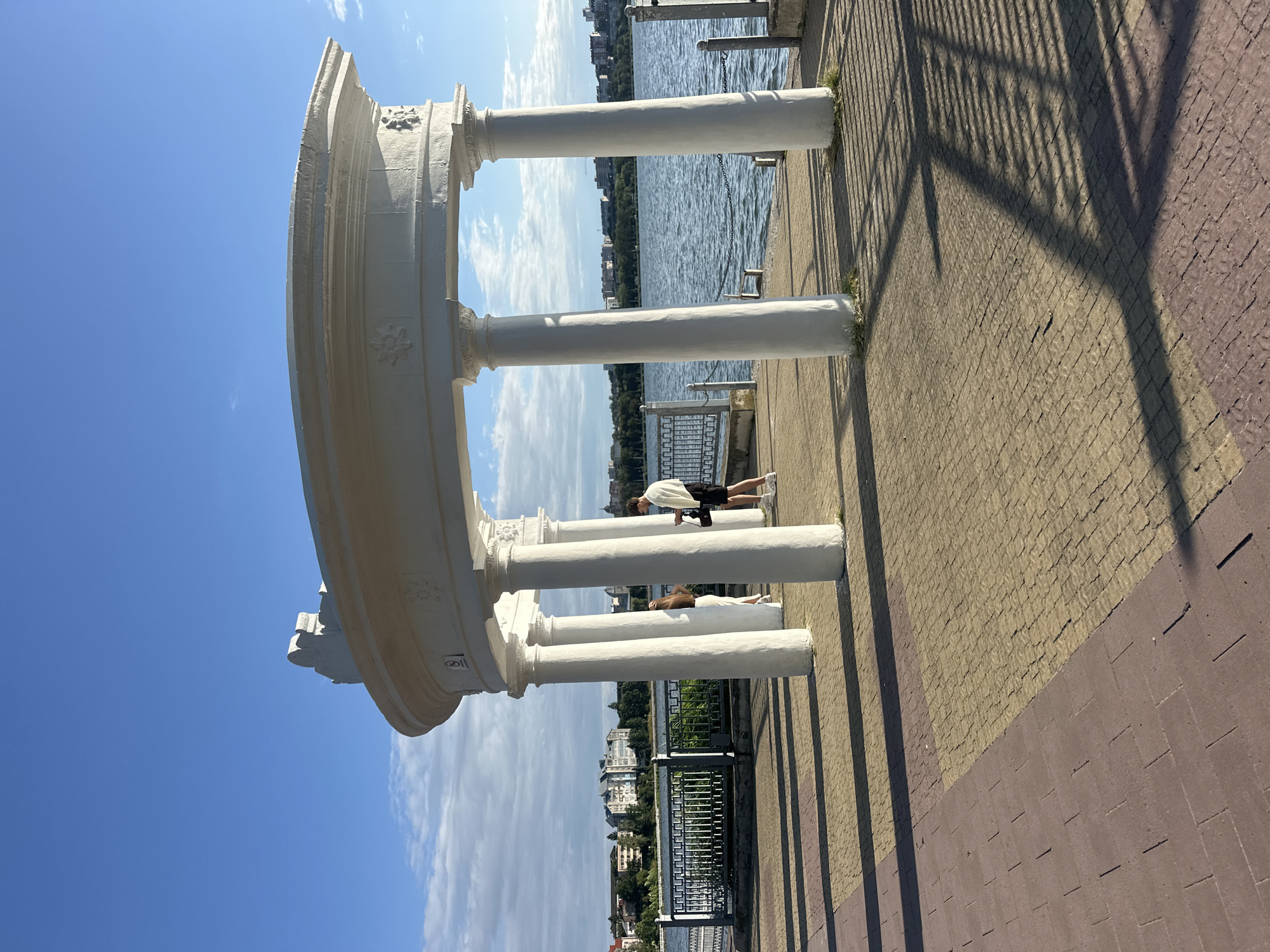 The image captures a picturesque scene at the Ternopil Pond in Ternopil, Ukraine. A white rotunda, an architectural structure with classical columns and ornate detailing, stands prominently in the foreground, casting long shadows on the patterned brick walkway. Two individuals are present: a woman in a white dress is partially visible near the left side of the rotunda, while a man in a black t-shirt and shorts stands to her right, holding a white garment over his shoulder. Beyond the rotunda, the calm waters of the pond stretch out, with a distant cityscape visible on the opposite bank, characterized by buildings and lush green trees. The sky is a clear, vibrant blue with a few wispy clouds. The lighting suggests it is either early morning or late afternoon, with the sun casting a warm glow and creating distinct shadows.