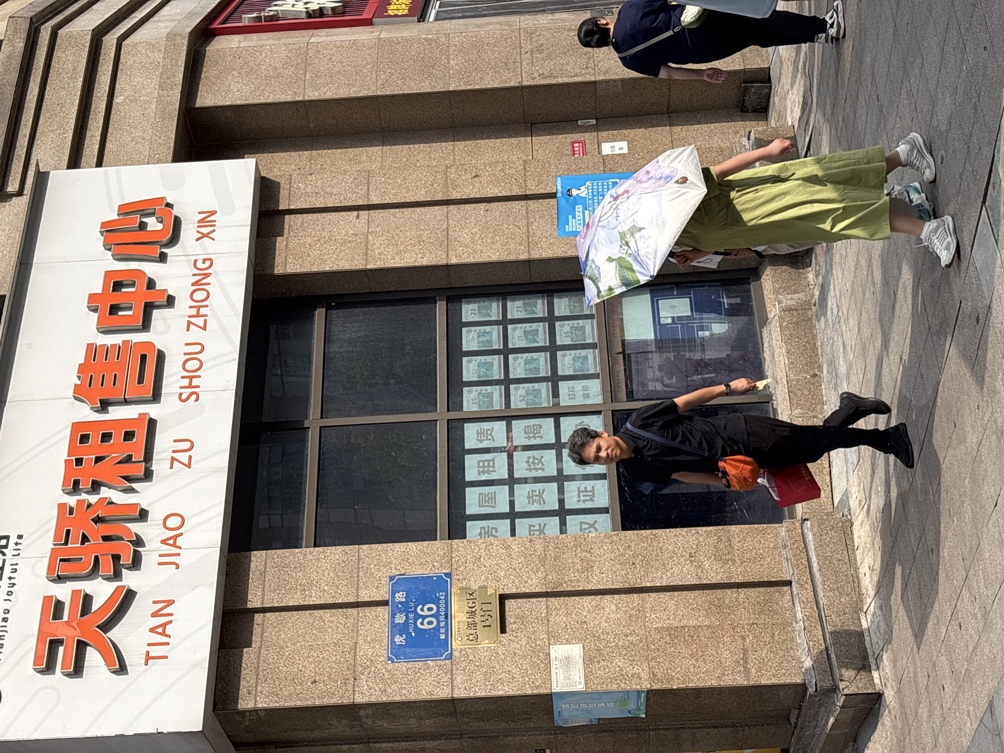 The image shows a street scene in the Yuzhong District of China. A building with a large sign displaying Chinese characters and English text reads "TIAN JIAO ZU SHOU ZHONG XIN" is visible. Below the main sign, a blue street sign indicates "HUXIE LU 66" and a postal code. Further down, a gold plaque reads "总部城 G区 1号门". Several people are walking on the sidewalk. One man in a black shirt and pants is carrying a red bag and an orange pouch. Another person, partially obscured by an umbrella, is wearing a green dress. A third person in a dark shirt and white pants walks away from the camera. Several posters are displayed in the windows of the building. The environment appears to be an urban setting with a paved sidewalk and the facade of a commercial building.