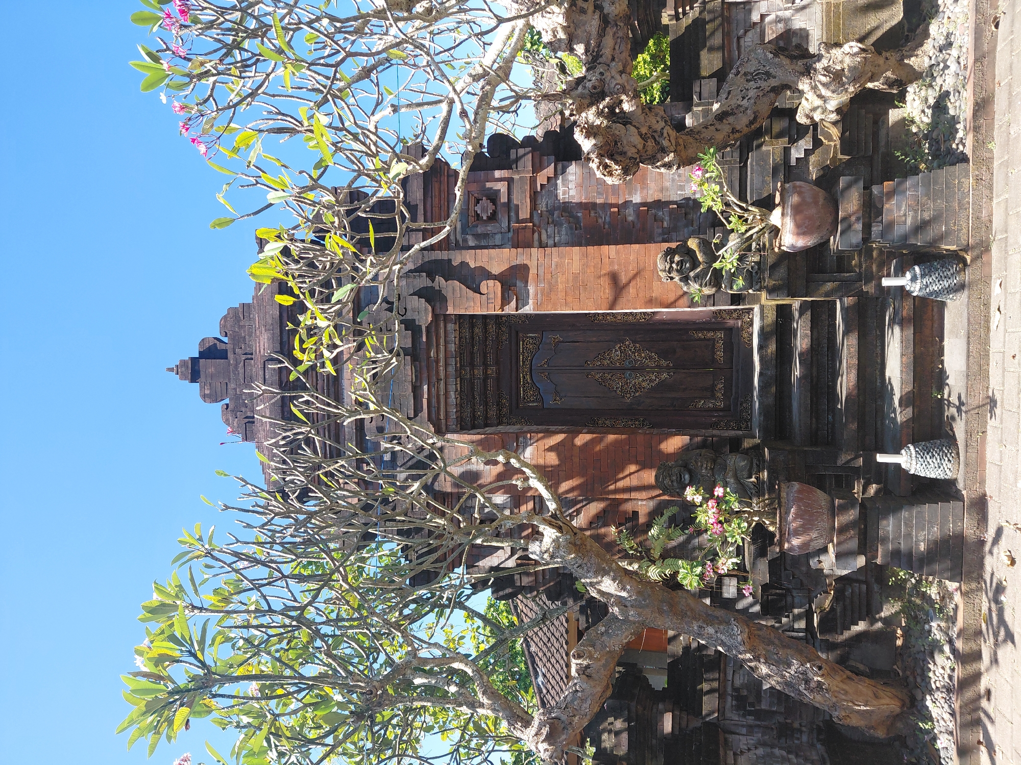 This is a photograph of the entrance to the Taman Sari Water Palace in Denpasar, Indonesia. The image is framed by two large frangipani trees, one on the left and one on the right, with bare branches and a few green leaves. In the center is a traditional Balinese temple gate made of dark brick. The gate has a carved wooden door with intricate gold detailing. Flanking the stairs leading to the door are two stone statues of seated figures. In front of the statues are potted plants with pink flowers. The sky above is a clear, bright blue. The ground in front of the gate is paved with stones and bricks, and there are shadows cast by the trees.