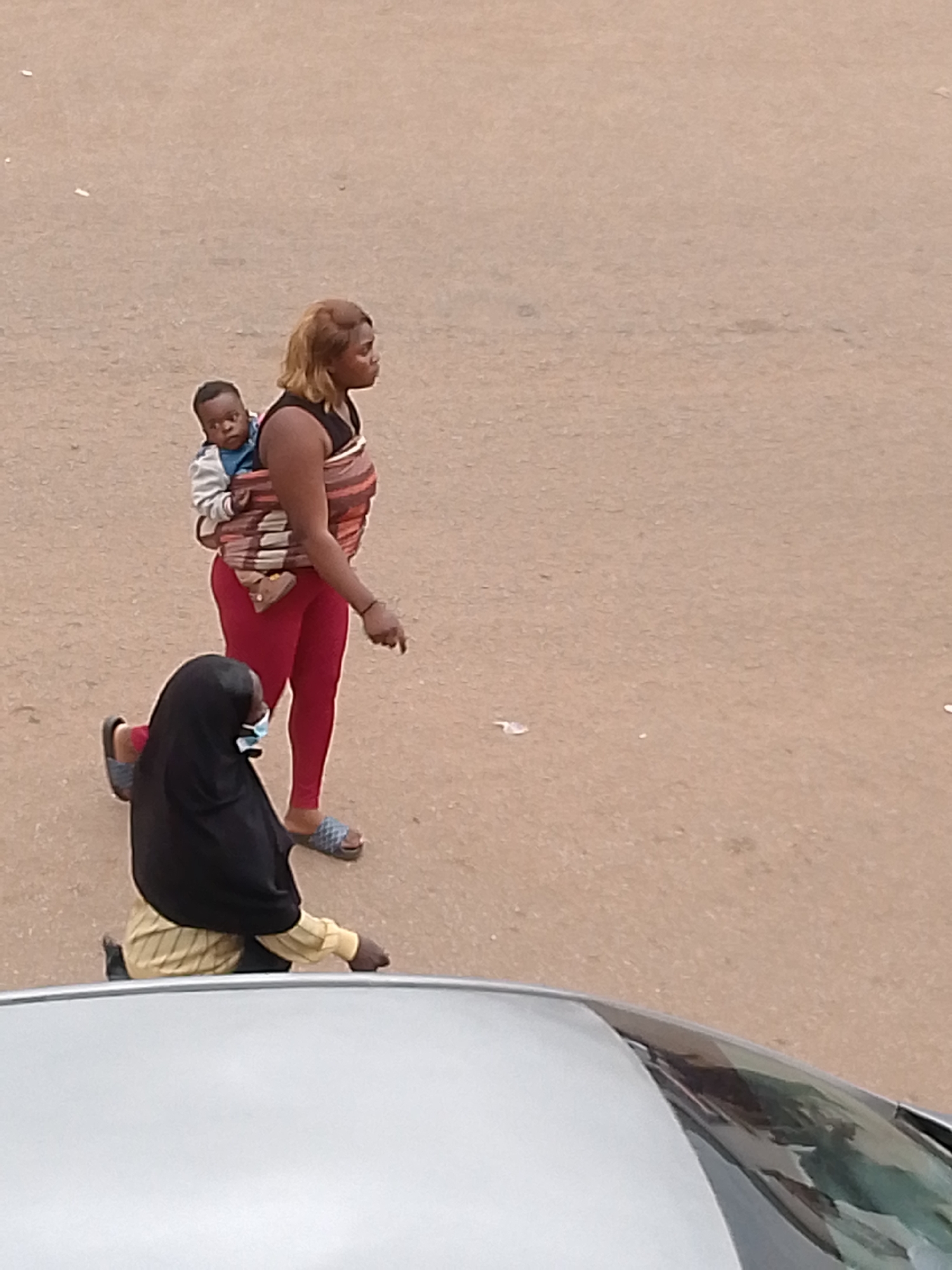 In Bukuru, Nigeria, an overhead shot captures two individuals on a dirt-colored, paved surface, possibly a road. The foreground is dominated by the top of a silver vehicle. To the left, a person wearing a black hijab and a light-colored patterned shirt is positioned low, with their legs bent, facing away from the viewer. They are also wearing a light blue face mask. To the right and slightly behind this individual, a woman with blonde hair walks away from the camera. She is carrying a baby on her back, secured by a multi-colored fabric wrap. The woman is wearing red leggings and a black sleeveless top. The environment appears to be an outdoor, possibly urban, setting with scattered small white debris on the ground.