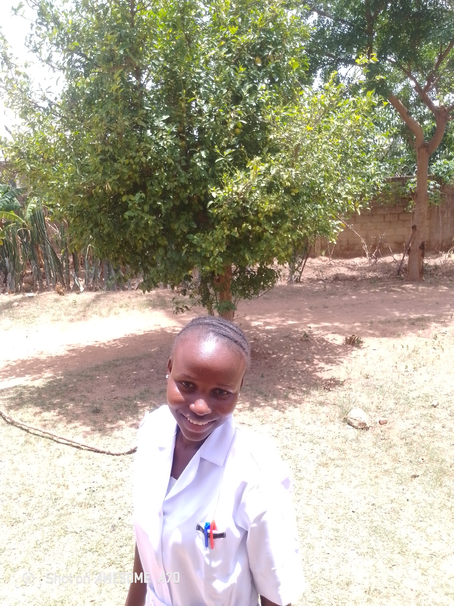 The image shows a young woman in a white lab coat standing outdoors in an area with sparse grass and sandy soil. She is smiling and looking directly at the camera. A small tree with dense green leaves is directly behind her. In the background, there is a tree and what appears to be a fence or wall. The location is in the city of Unknown, Unknown.