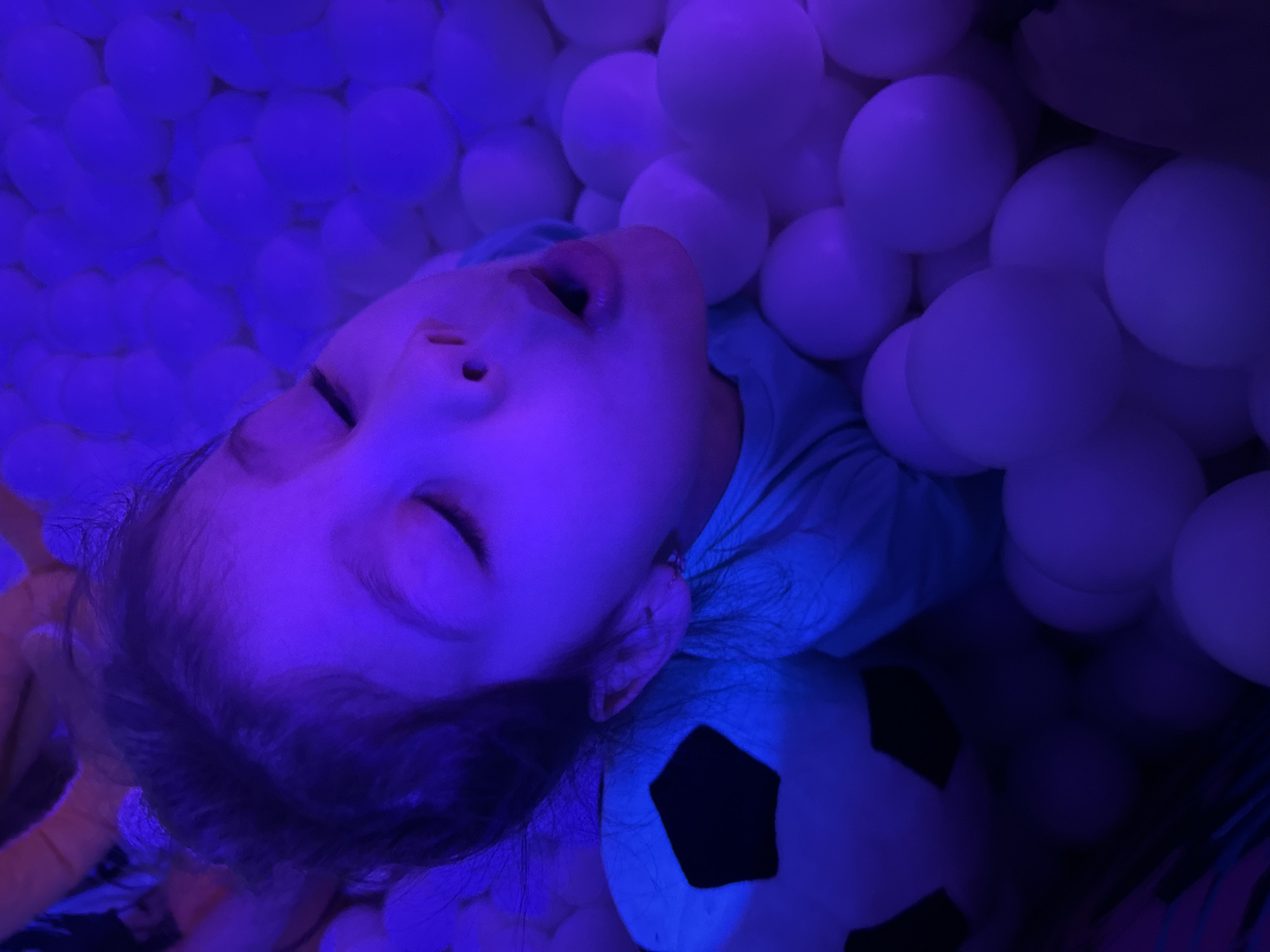 A young child is depicted lying on their back, deeply immersed within a ball pit, in the city of Unknown, Unknown. The child's eyes are closed, and their mouth is slightly agape. Dark hair is visible, parted on the head. The child wears a light-colored t-shirt, appearing teal or light blue, which is illuminated by the ambient light. Surrounding the child are numerous white spherical plastic balls, filling the frame. To the left of the child's head, a plush soccer ball is partially visible, featuring white panels that glow under the lighting and dark, likely black, pentagonal patches. The entire scene is bathed in an intense, artificial purple-blue light, causing the child's skin and the white balls to appear with a strong purplish hue. The lighting suggests a controlled or specialized environment, possibly a play area or sensory room.