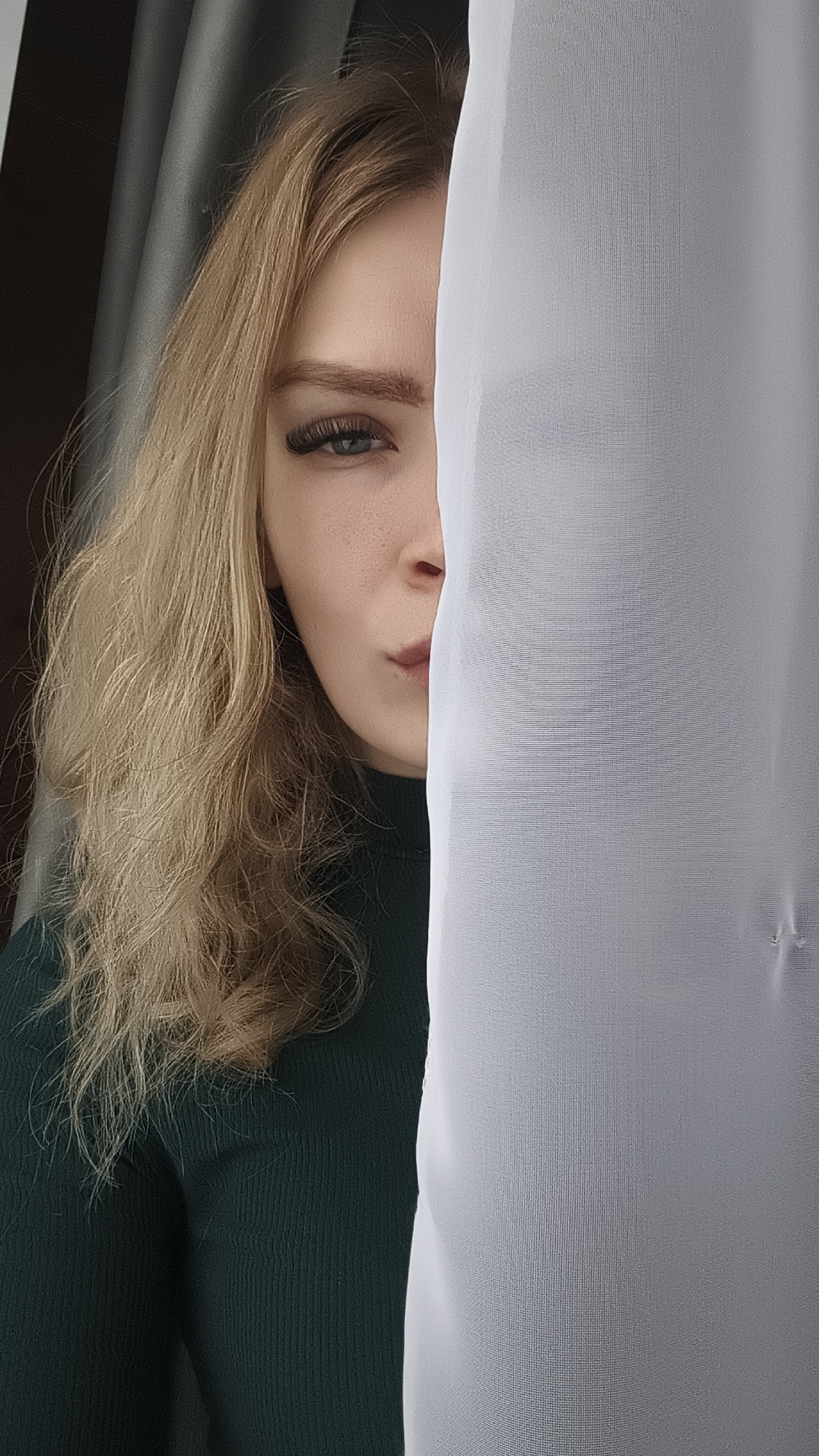 A fair-skinned young woman is partially visible, obscured by a sheer white curtain. Only half of her face is exposed, revealing a defined eyebrow, eyelashes, and a portion of her mouth. She is wearing a dark green turtleneck sweater. Her blonde hair is wavy and falls to her shoulder. The background includes another curtain or wall in a dark color. This photograph does not provide enough information to determine a specific location in Русский Вожой, Russia.