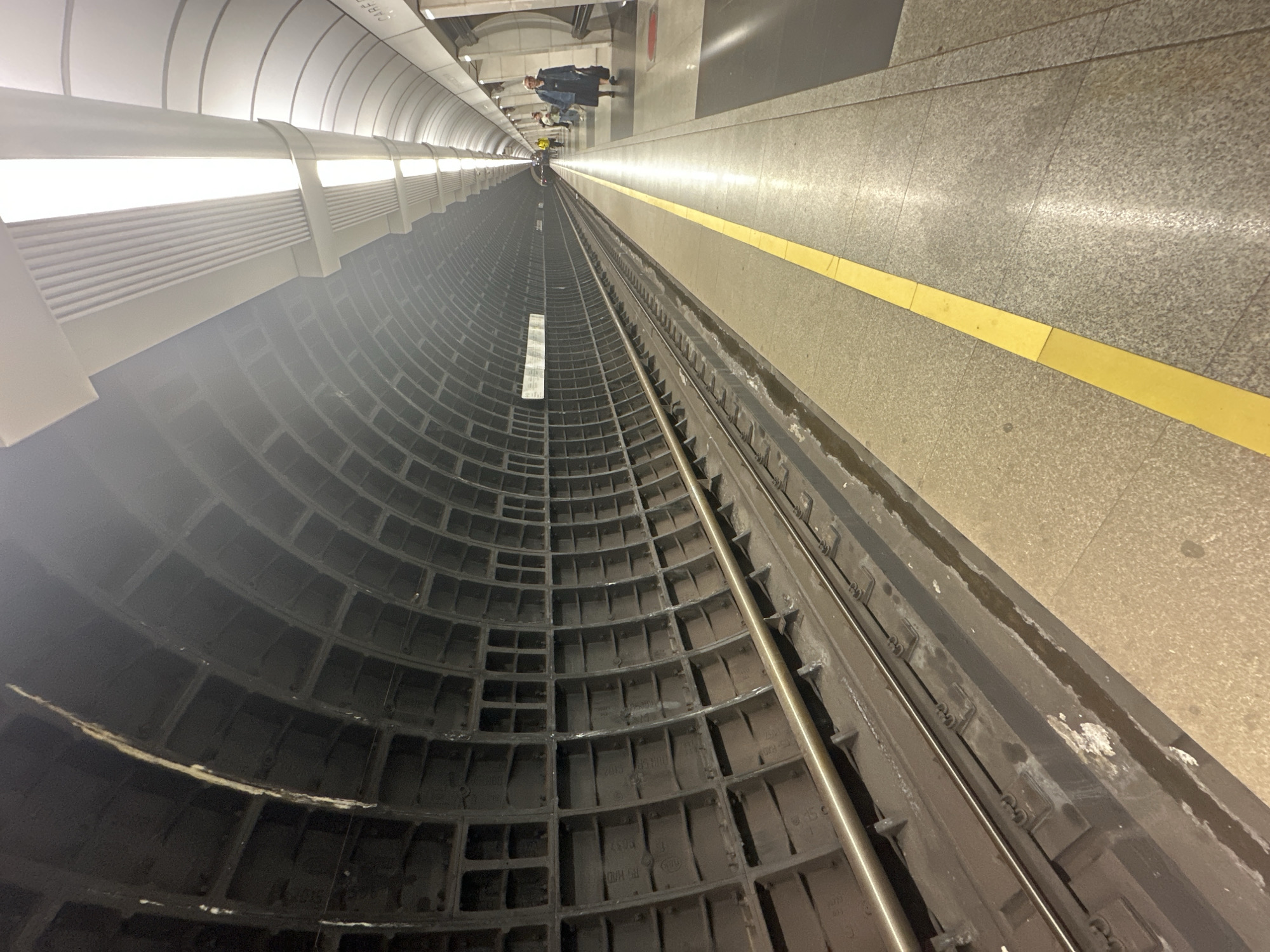 The image shows a metro station platform in Moscow, Russia. The platform is on the right side of the image, constructed of granite tiles with a yellow tactile strip indicating the platform edge. Metro tracks run along the platform's edge. The tunnel wall on the left is made of concrete blocks arranged in a honeycomb pattern, lit by linear lights installed above. A small group of people is visible in the distance, standing on the platform.