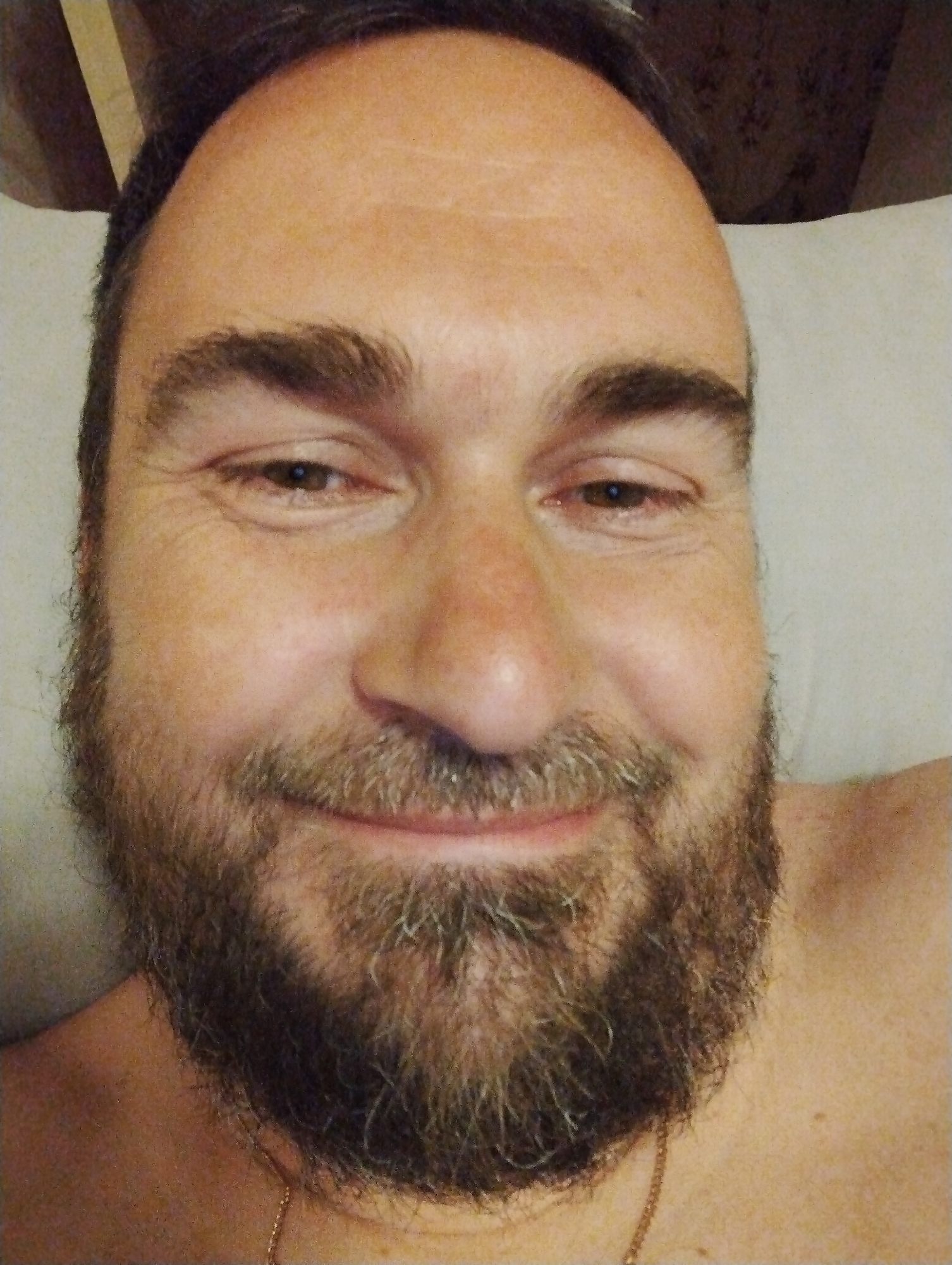 A close-up, head-and-shoulders selfie of a man with a beard is shown. He is looking directly at the camera with a slight smile. The man has brown hair and light brown eyes. He is wearing a gold chain necklace. The background is out of focus and appears to be a light-colored surface, possibly a pillow or wall. The man's skin tone is fair, and there are some wrinkles visible around his eyes. The image is taken in low light conditions, which gives it a soft, diffused quality.