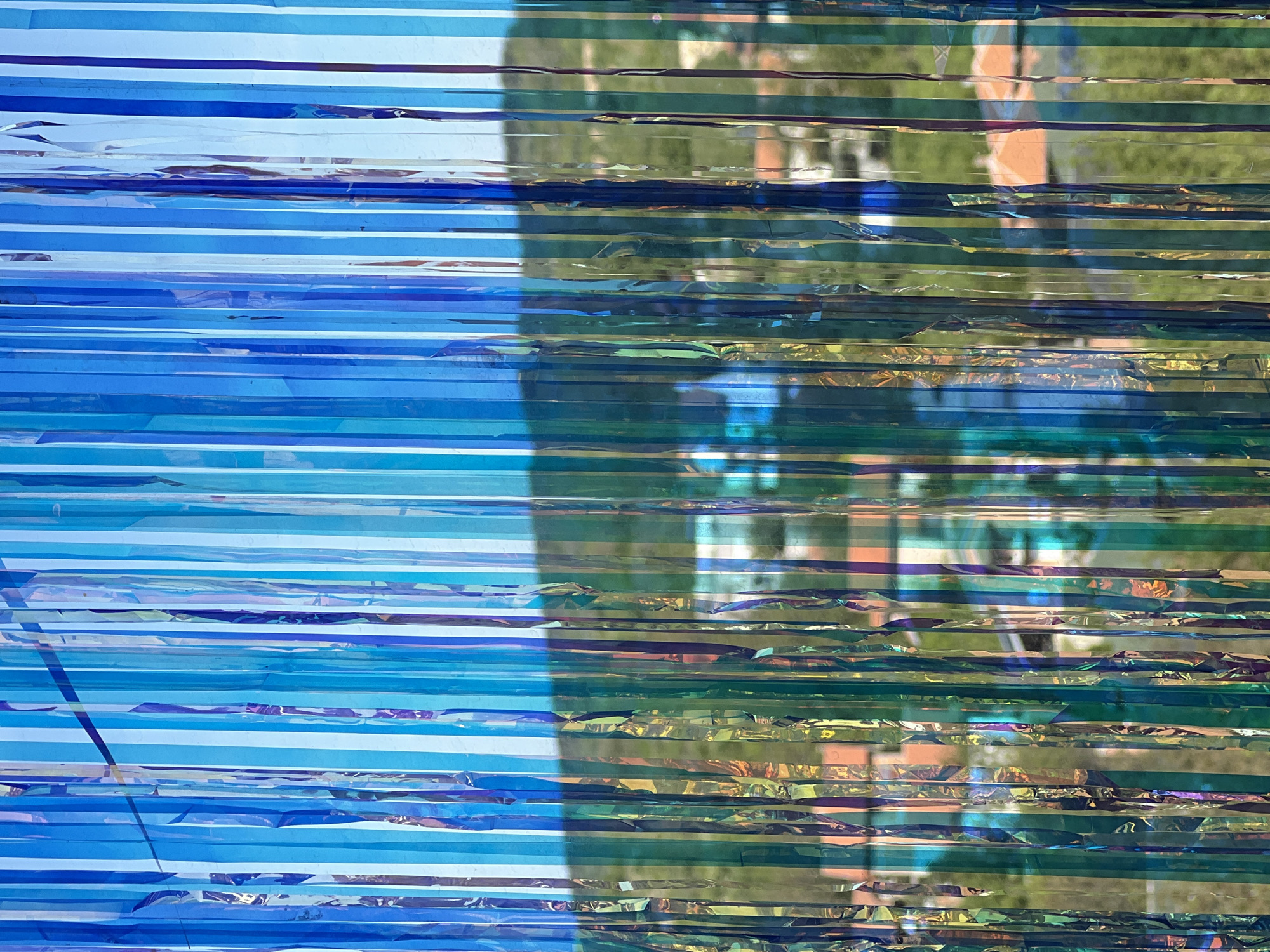 The image depicts a set of thin, horizontal streamers in shades of blue on the left and a mix of reflective blue, green, and gold on the right. The background visible through the streamers is blurry, suggesting a scene with trees and buildings. The location is Palilula Urban Municipality, Serbia, but specific details about the people, subjects, objects, actions, or interactions are not clearly discernible from the image. The focus is on the texture and color of the streamers and the distorted view of the background.