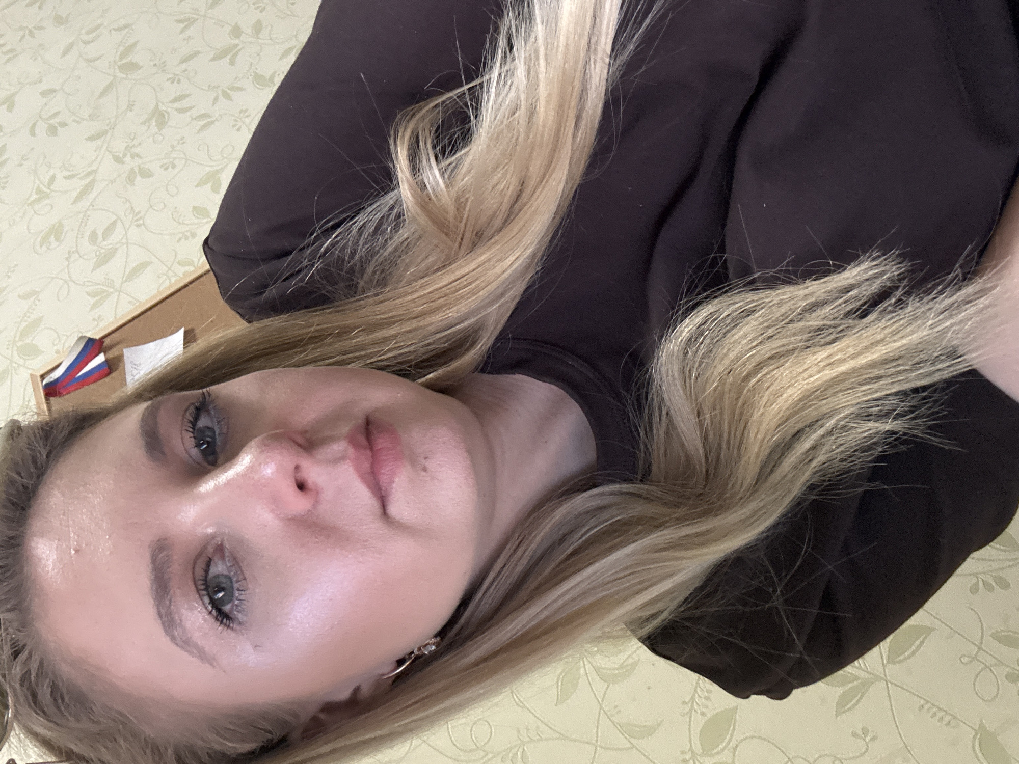 This is a close-up, sideways selfie of a young woman with blonde hair. She is wearing a dark, long-sleeved top. Her hair is long and appears to be slightly wavy. Her face is smooth and appears to have makeup on, including mascara and possibly lipstick. She is wearing a small hoop earring with what looks like a few small stones. Her expression is neutral, and the lighting suggests it might be indoors, possibly during the day. The background is a light-colored wall with a faint floral pattern. To the left of the woman's head, a small object with red, white, and blue stripes is visible, possibly a flag or a ribbon. The overall scene is simple and intimate, likely a personal photograph. There is no discernible text in the image.