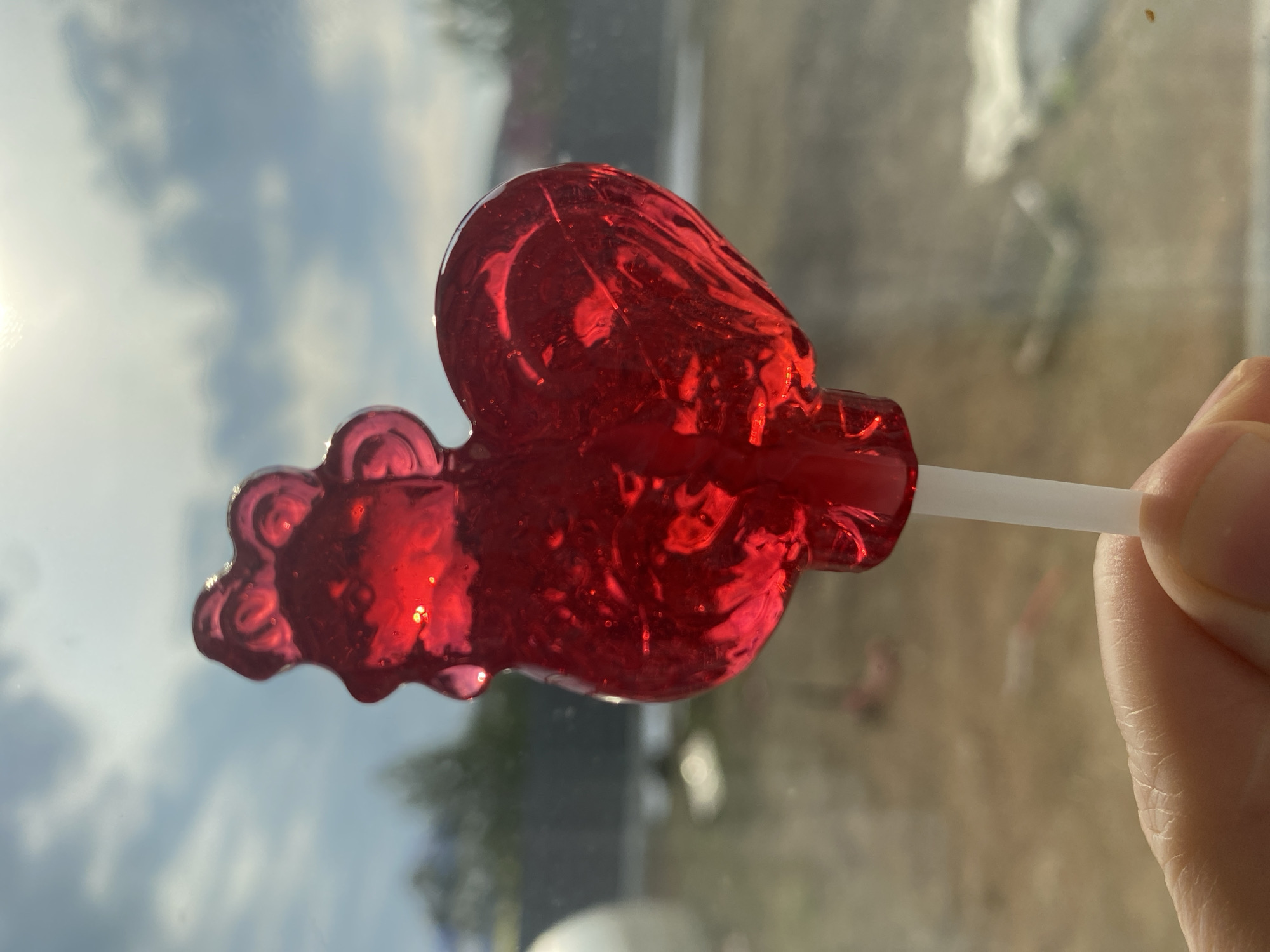 A red, heart-shaped lollipop on a white stick is being held in front of a window. The background visible through the window suggests an outdoor scene with sky and trees. The location is not explicitly evident from the image, so providing the location as Chaikovsky, Russia would be speculative.
