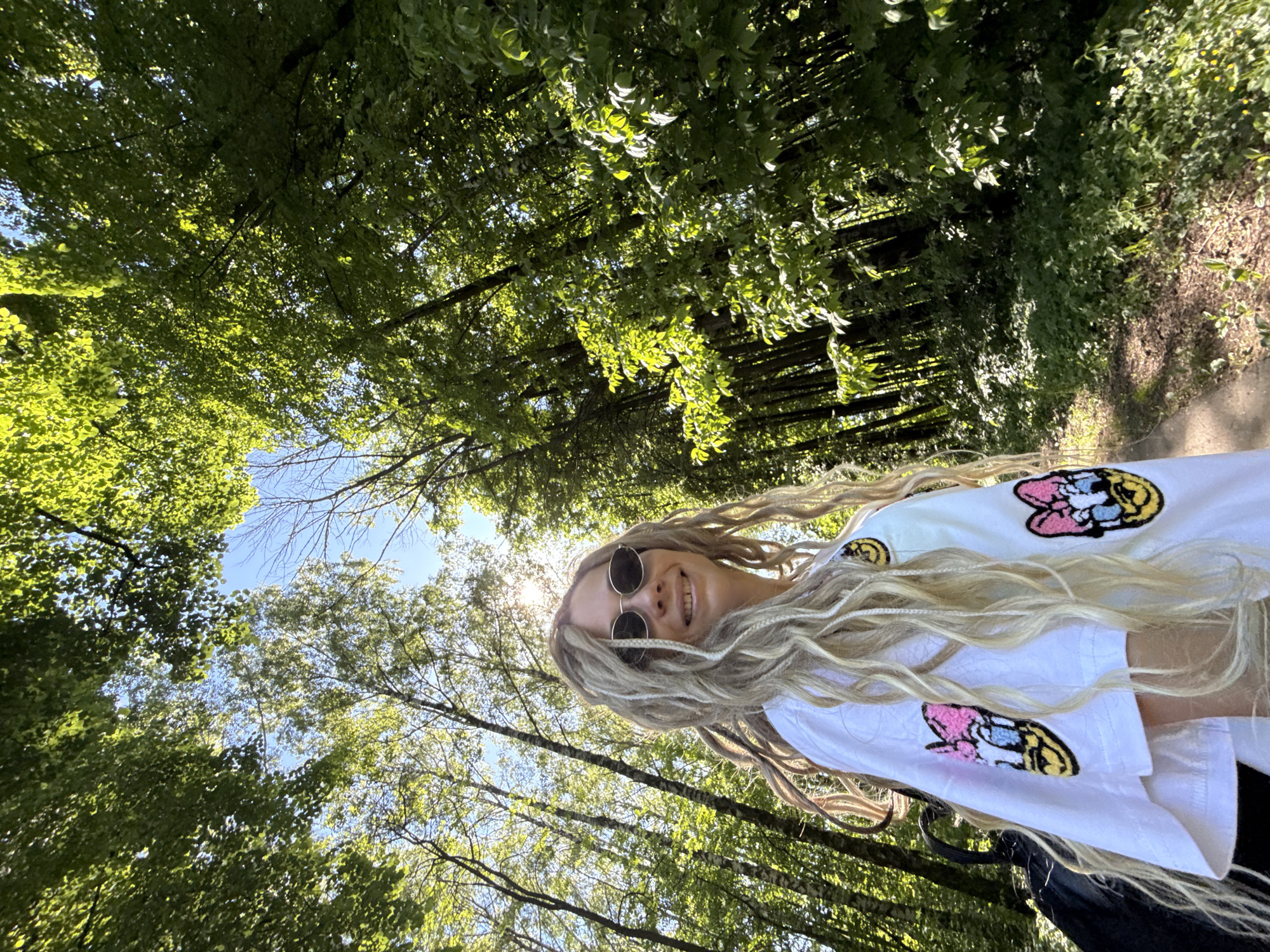This image, captured in Rusanovo, Russia, displays a female individual in a sunlit wooded environment. The woman is positioned in the foreground, viewed from a low angle, with long blonde hair styled in dreadlocks. She wears dark, oval-shaped sunglasses and a white short-sleeved shirt adorned with at least two embroidered patches featuring the character Daisy Duck on the visible right side of the garment. A dark backpack is visible behind her left shoulder. Her facial expression indicates a smile, directed towards the camera. The background consists of a dense forest of numerous tall deciduous trees with vibrant green foliage. Sections of clear blue sky are visible through gaps in the tree canopy. Direct sunlight filters through the leaves, creating bright highlights and dappled shadows on the foliage and ground. A direct solar glare point is present in the upper right quadrant of the frame. A narrow dirt path is visible in the lower right section, bordered by dense green undergrowth and small plants.